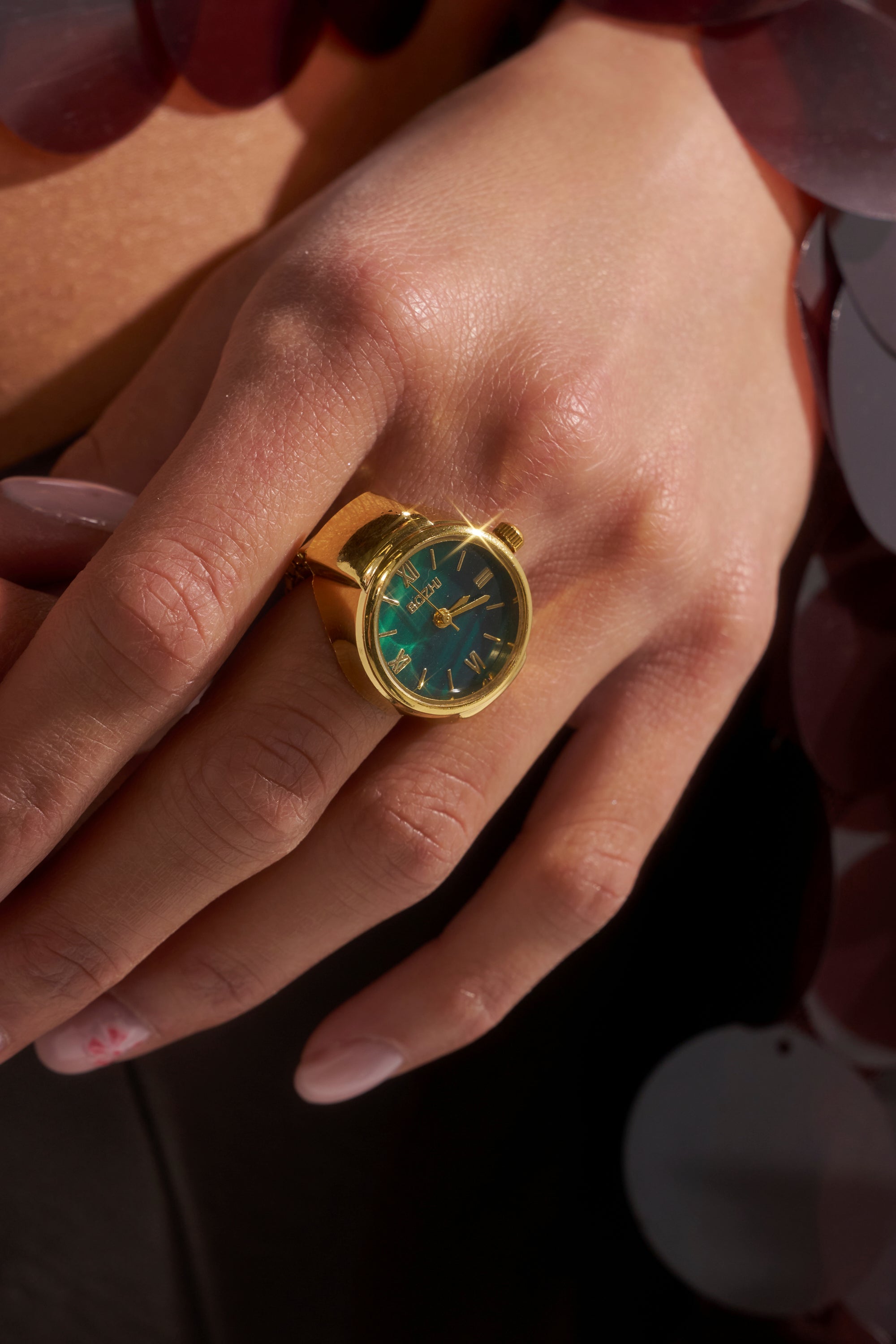 A hand wears the WHAT TIME IS IT RING—a gold metallic watch ring styled like a mini wristwatch with a green face and gold hour markers. Manicured nails have light polish, with a subtle design accent on one nail.