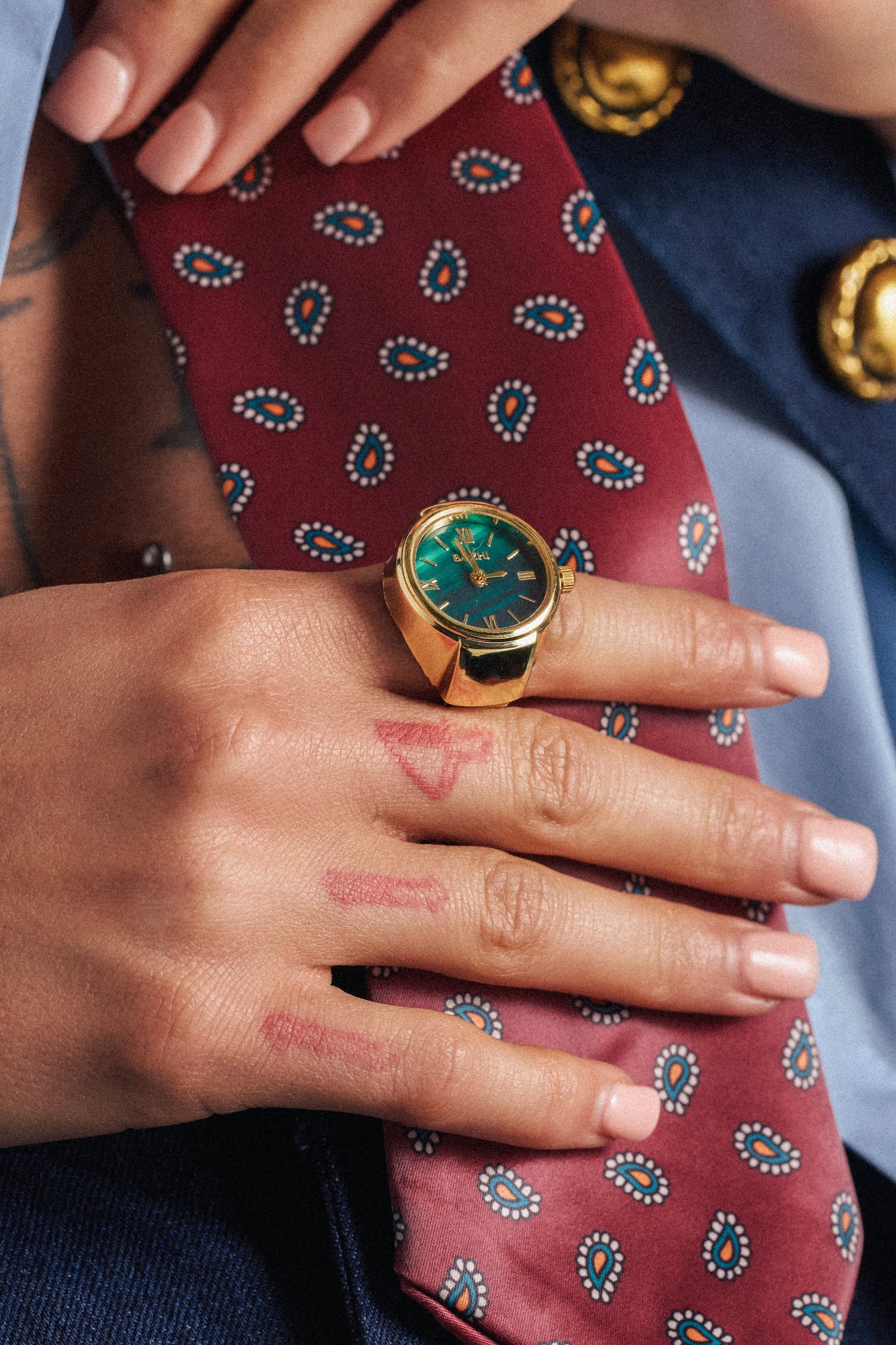 A hand with manicured nails and lipstick marks rests on a red paisley tie, wearing the striking WHAT TIME IS IT RING—a gold metallic watch ring with a green face—while blue clothing with gold buttons is partially visible. 0