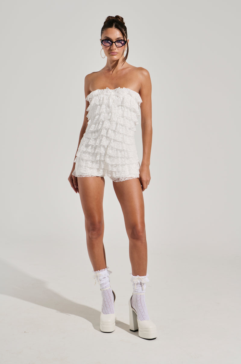 A person stands against a plain, light-colored background wearing the DESERT TIME ROMPER, characterized by its layered lace ruffles and a stretchy inner lining. They complete the look with white lace socks, white chunky platform shoes, large hoop earrings, glasses, and an updo hairstyle.