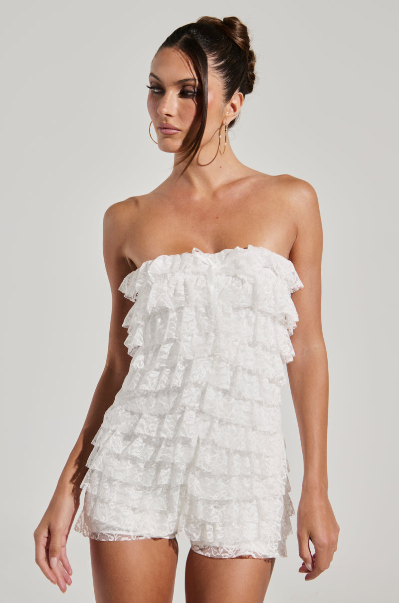 A woman poses gracefully against a plain backdrop, showcasing the DESERT TIME ROMPER. The white strapless romper is adorned with multiple layers of lace ruffles and offers a stretchy inner lining for added comfort. Her hair is styled in a neat bun and she accessorizes with large hoop earrings, complementing the chic ensemble.