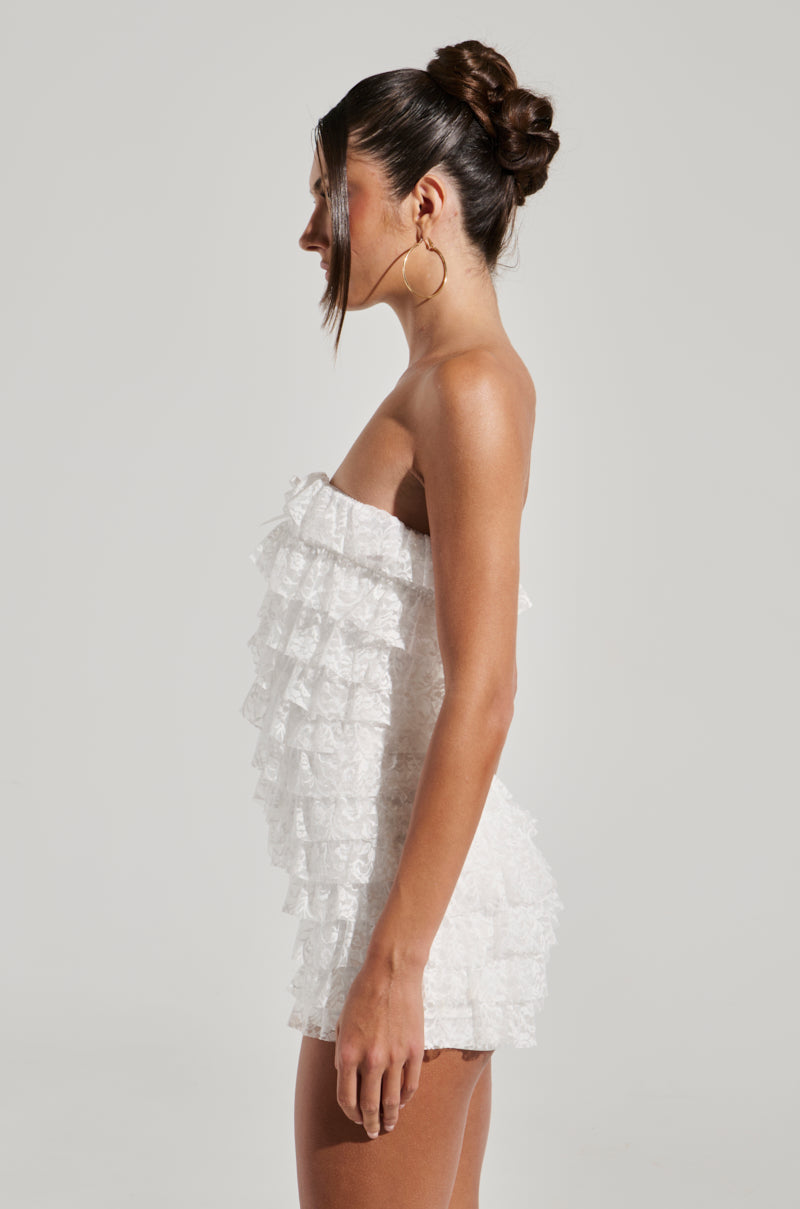 A woman with a bun hairstyle stands in side profile against a plain background, wearing the DESERT TIME ROMPER—a strapless, short, white dress adorned with multiple layers of lace ruffles and featuring a stretchy inner lining. She has large hoop earrings and appears relaxed.