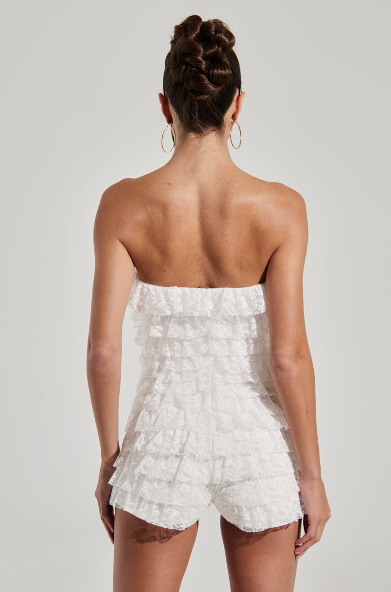 A person with a braided updo is wearing a white, strapless DESERT TIME ROMPER adorned with lace ruffles. The photo, taken from the back, highlights the layers of ruffles on the garment and showcases their hoop earrings. The romper's stretchy inner lining ensures comfort while the light-colored background complements the outfit beautifully.