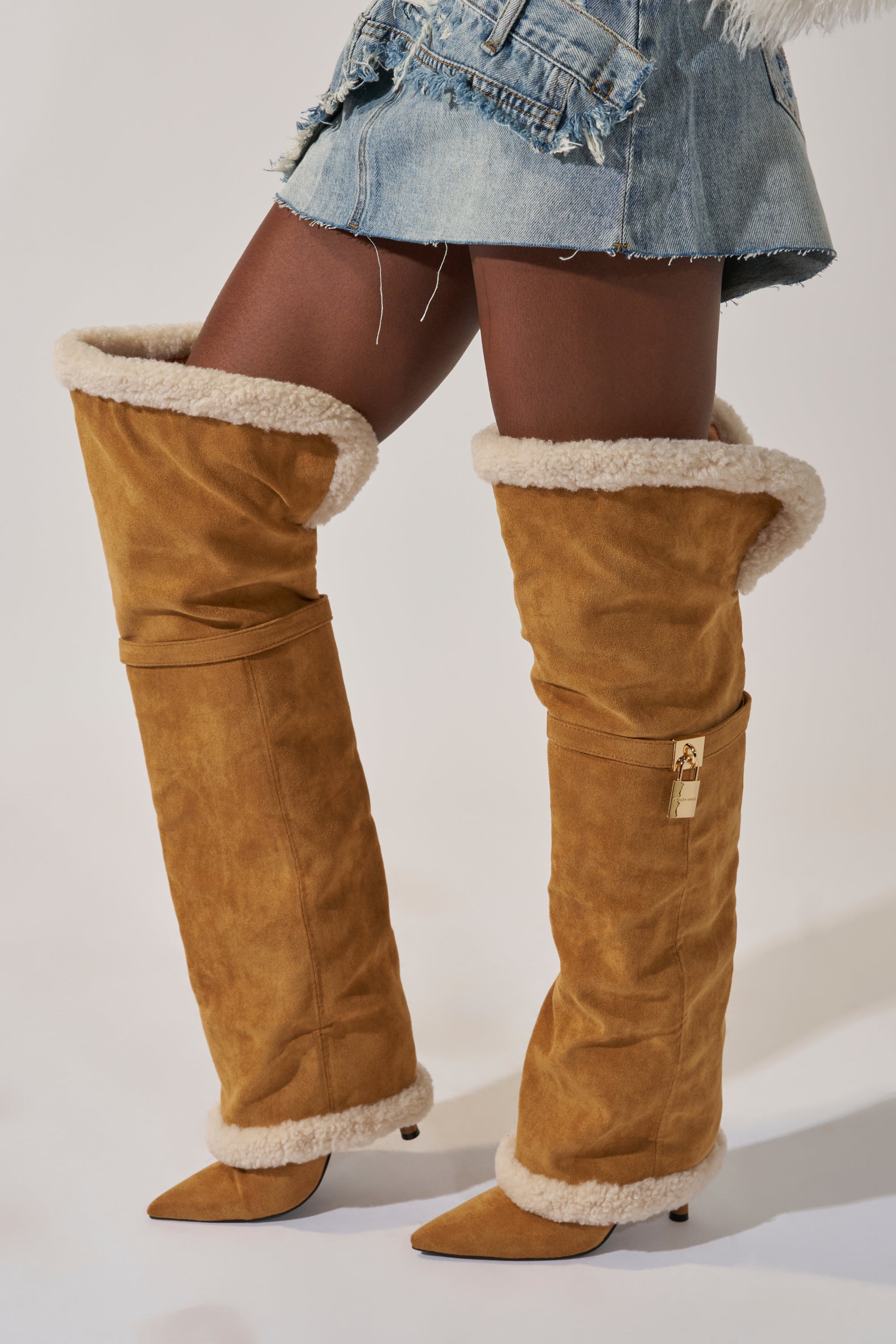A person wearing an asymmetrical denim mini skirt and oversized tan AZALEA WANG RACHANA TAN SHERPA STILETTO BOOTS with pointed toes and a small gold lock detail.