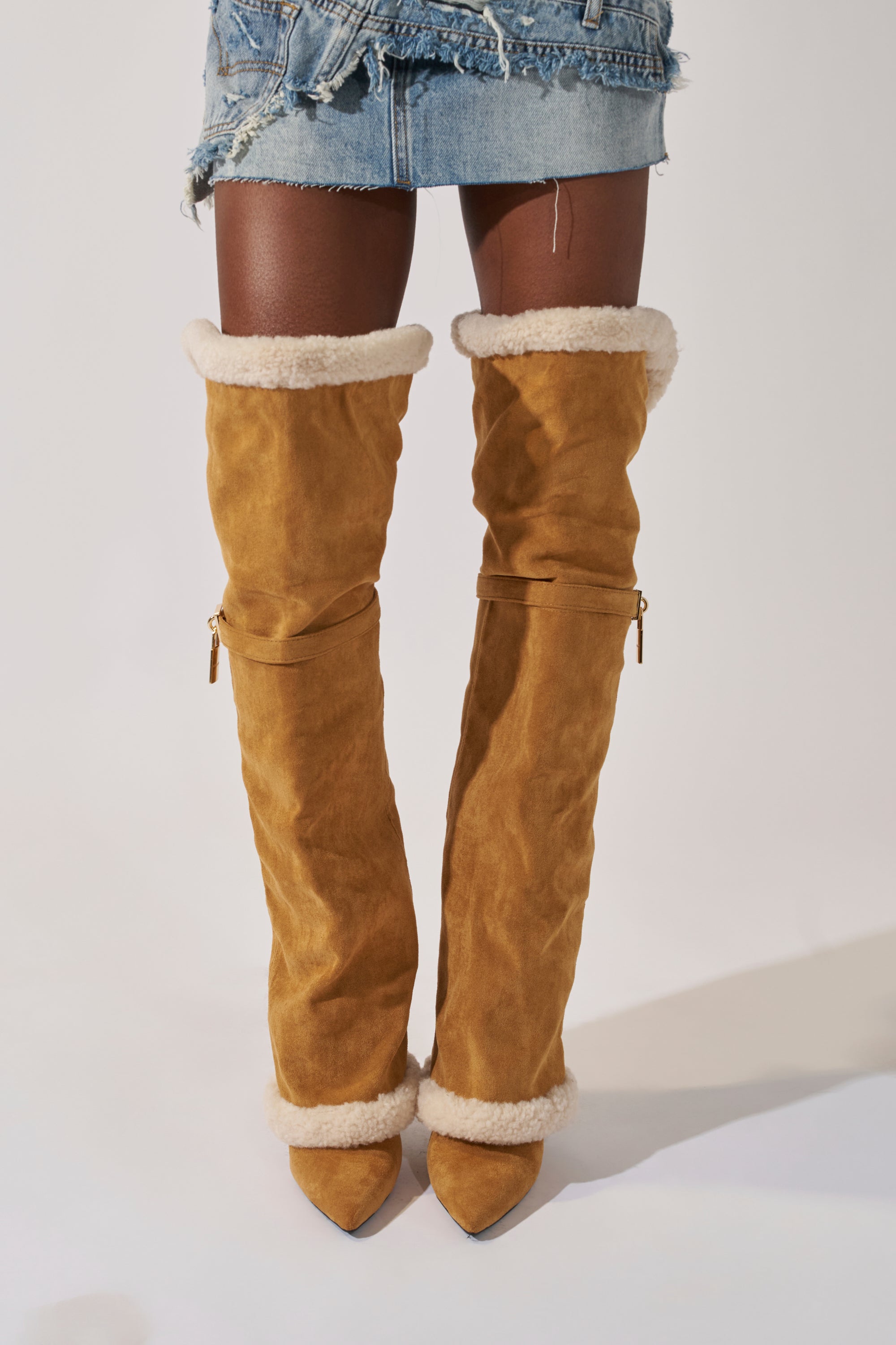 A person models the AZALEA WANG RACHANA TAN SHERPA STILETTO BOOT, styled with a distressed denim mini skirt. Only the legs and skirt are shown against a plain background.