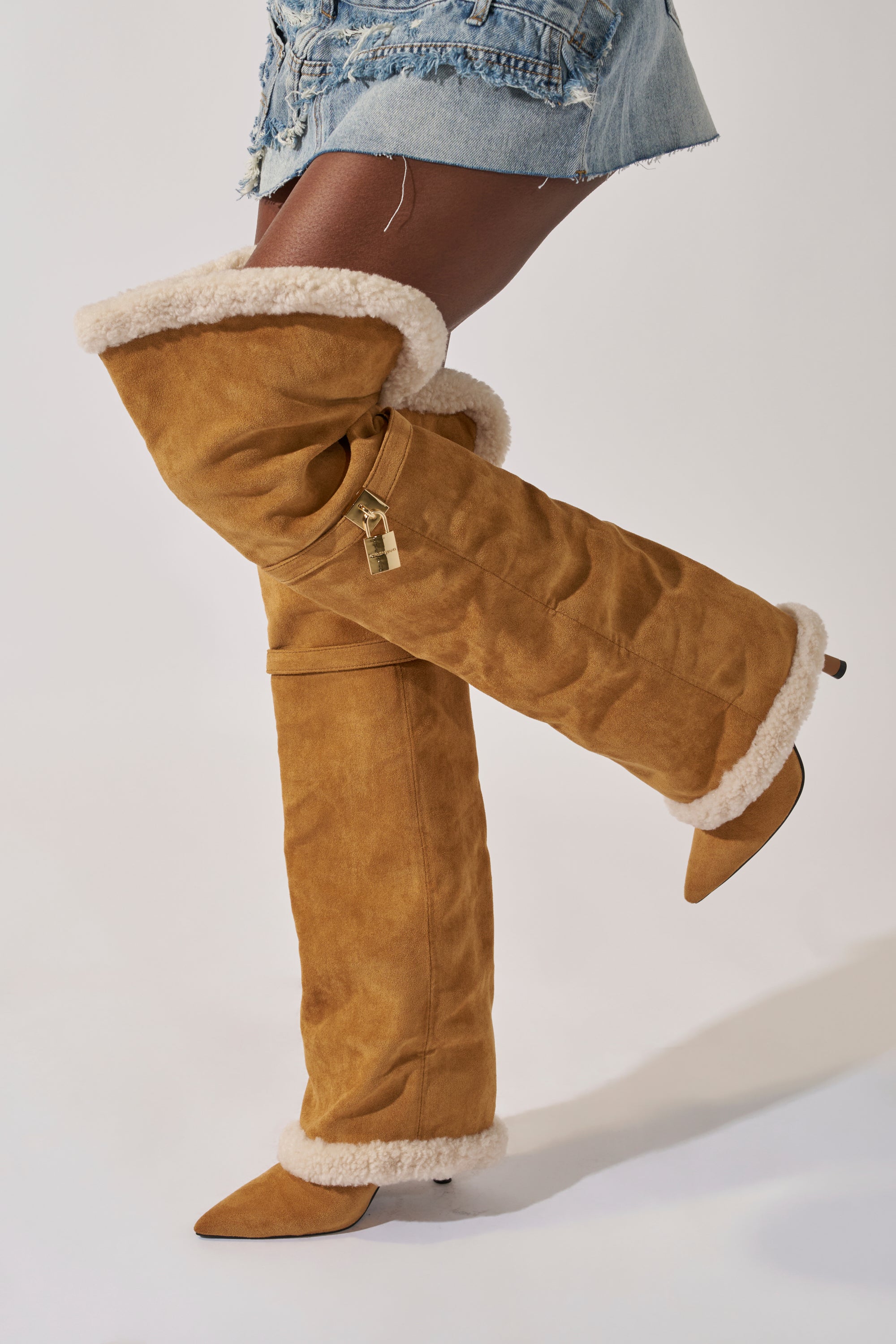 A person models the AZALEA WANG RACHANA TAN SHERPA STILETTO BOOT—tan, fur-lined, knee-high boots with pointed toes and a lock charm—styled with frayed denim shorts against a plain background.