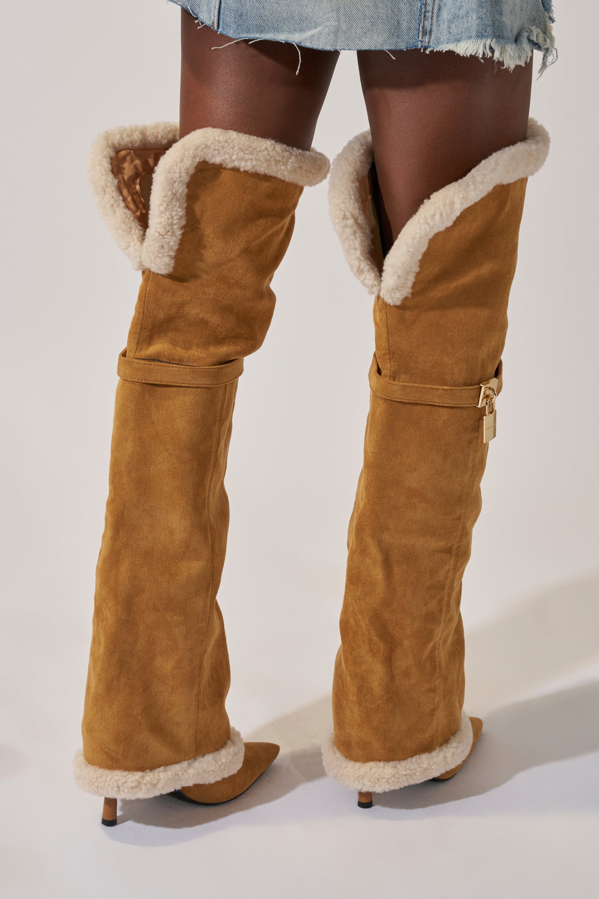 A person wears the AZALEA WANG RACHANA TAN SHERPA STILETTO BOOT—tan suede, knee-high with white shearling trim, pointed stiletto heels, and a relaxed slouchy fit—paired with a frayed denim mini skirt.