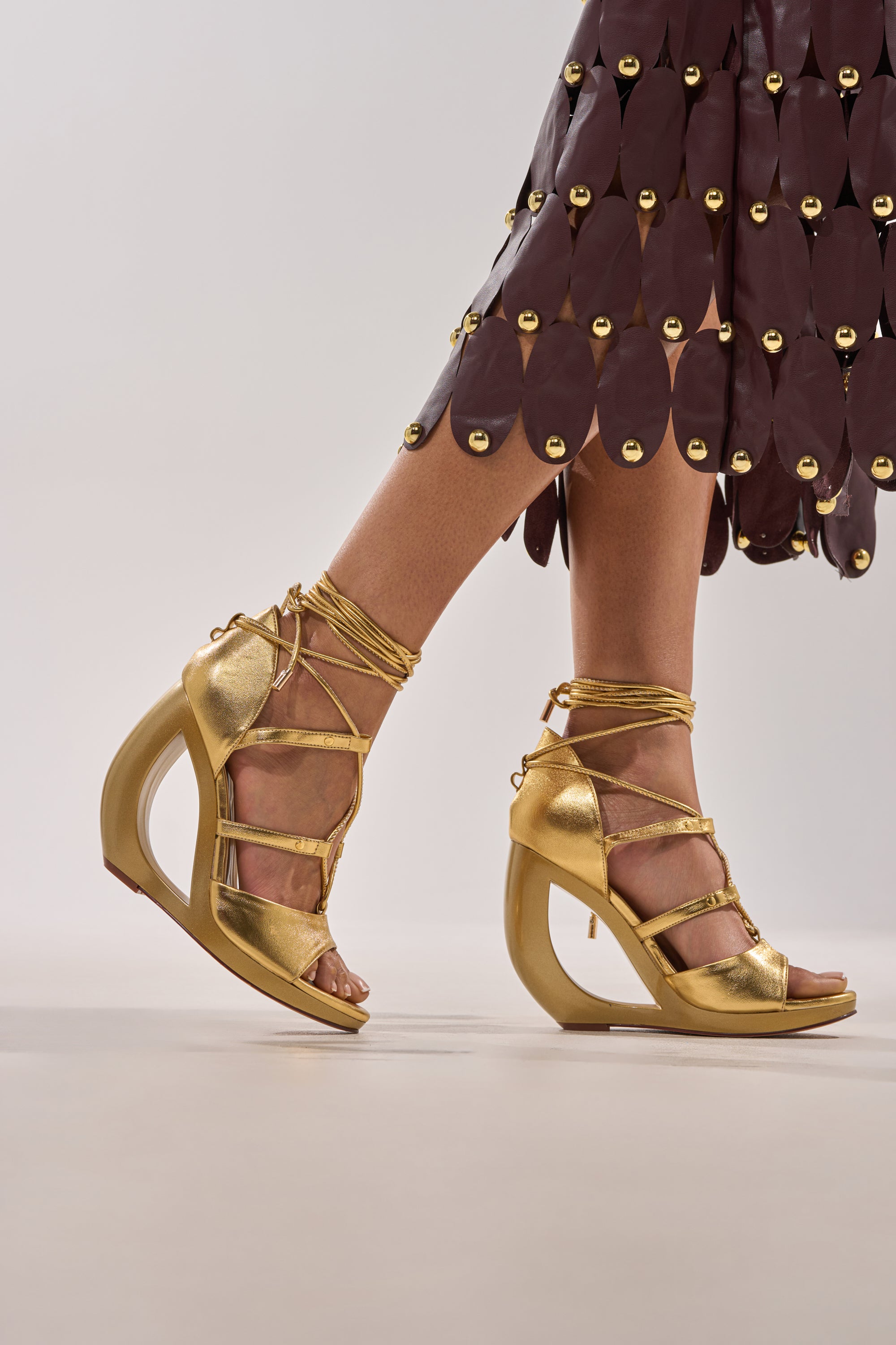 Wearing AKIRA GOLD RACHNA GOLD WEDGE OPEN TOE SANDAL with comfort cushion insole and ankle straps, styled with a brown skirt adorned with large oval embellishments and gold studs. 1