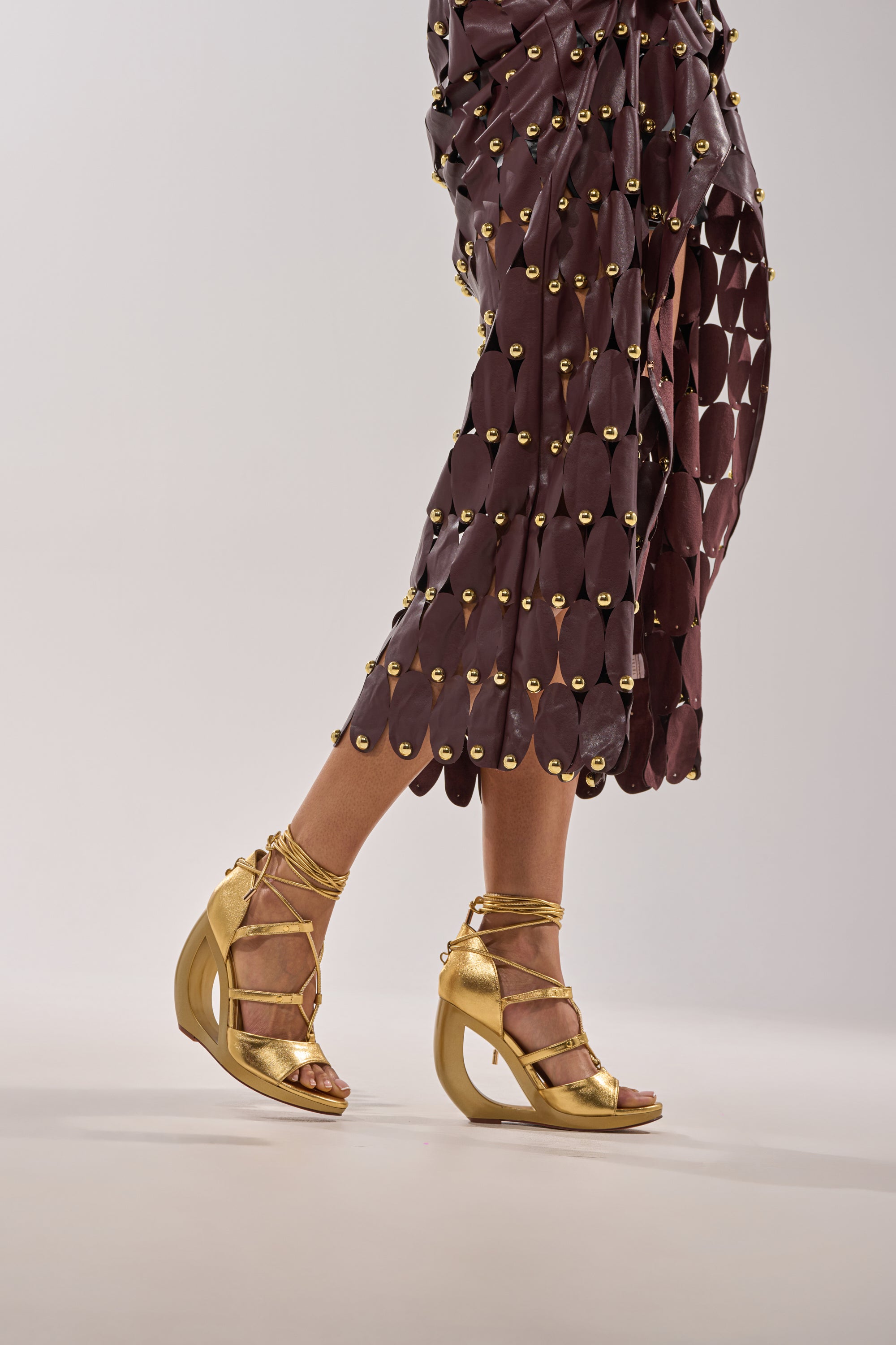 Wearing a brown geometric-patterned skirt with gold studs and AKIRA GOLD RACHNA GOLD WEDGE OPEN TOE SANDALS with crisscross ankle straps, the person stands against a light neutral background. 2