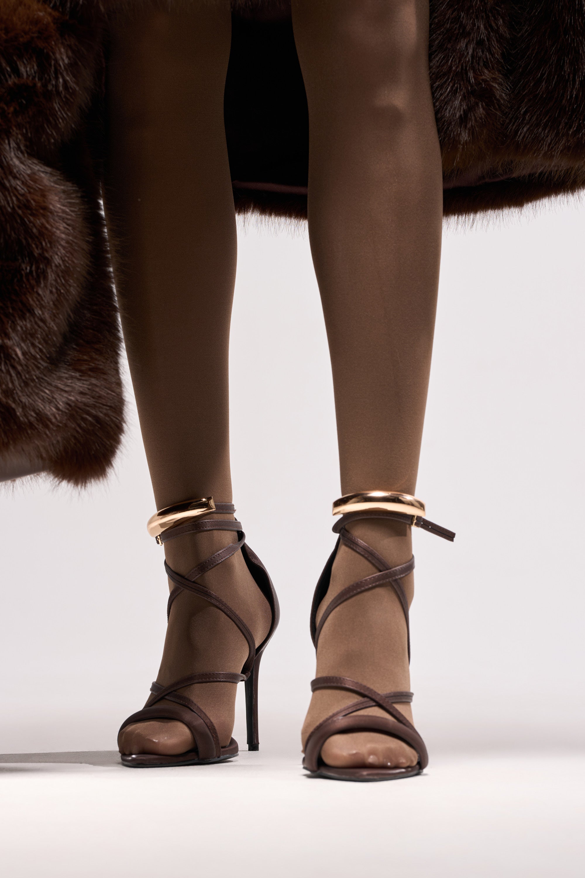 Wearing sheer brown tights, gold anklets, and the AZALEA WANG RADHA CHOCOLATE METAL EMBELLISHED SANDAL, a person stands with a brown fur coat draped above their knees against a plain light background. 1