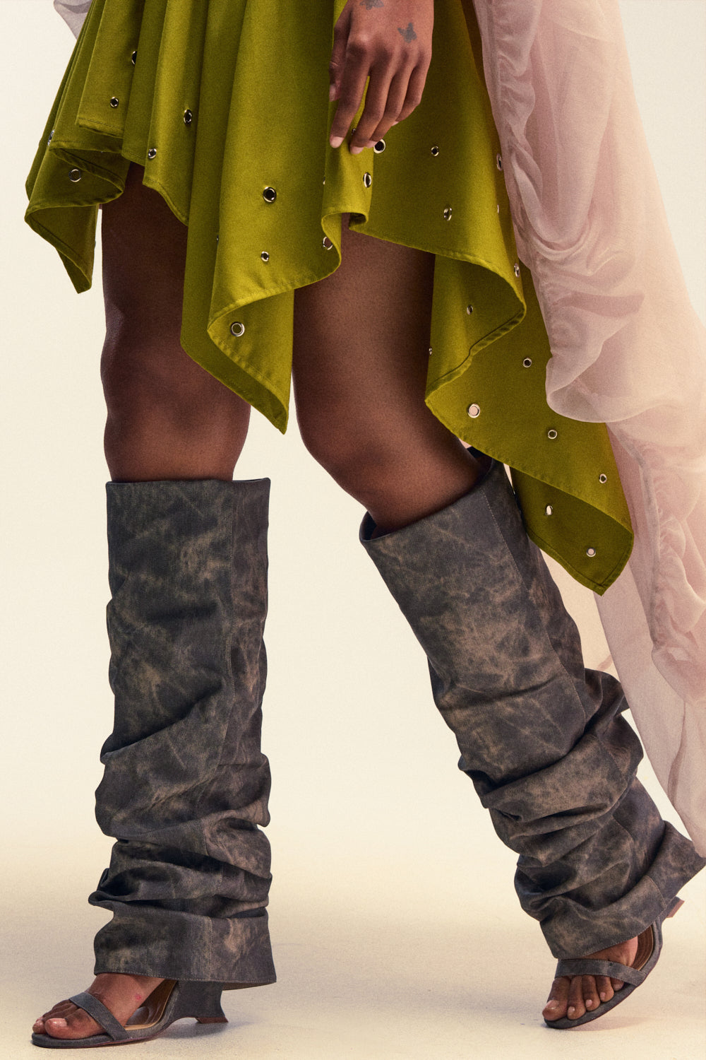 Wearing a green skirt with metal grommets, a sheer pink drape, and brown slouchy leg warmers that emphasize a relaxed statement silhouette, the individual completes the look with AZALEA WANG BROWN ACID WASHED DENIM GLADIATOR SANDALS while walking forward.