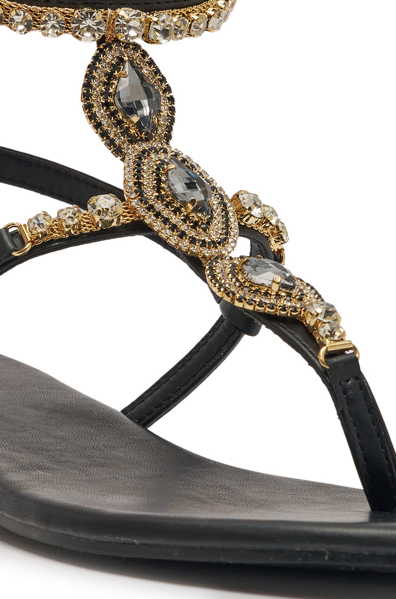 Close-up of the AZALEA WANG RAFEE BLACK RHINESTONE EMBELLISHED FLAT STRAP SANDAL in faux leather, featuring gold and clear gemstone accents with intricate decorative straps that sparkle. 7