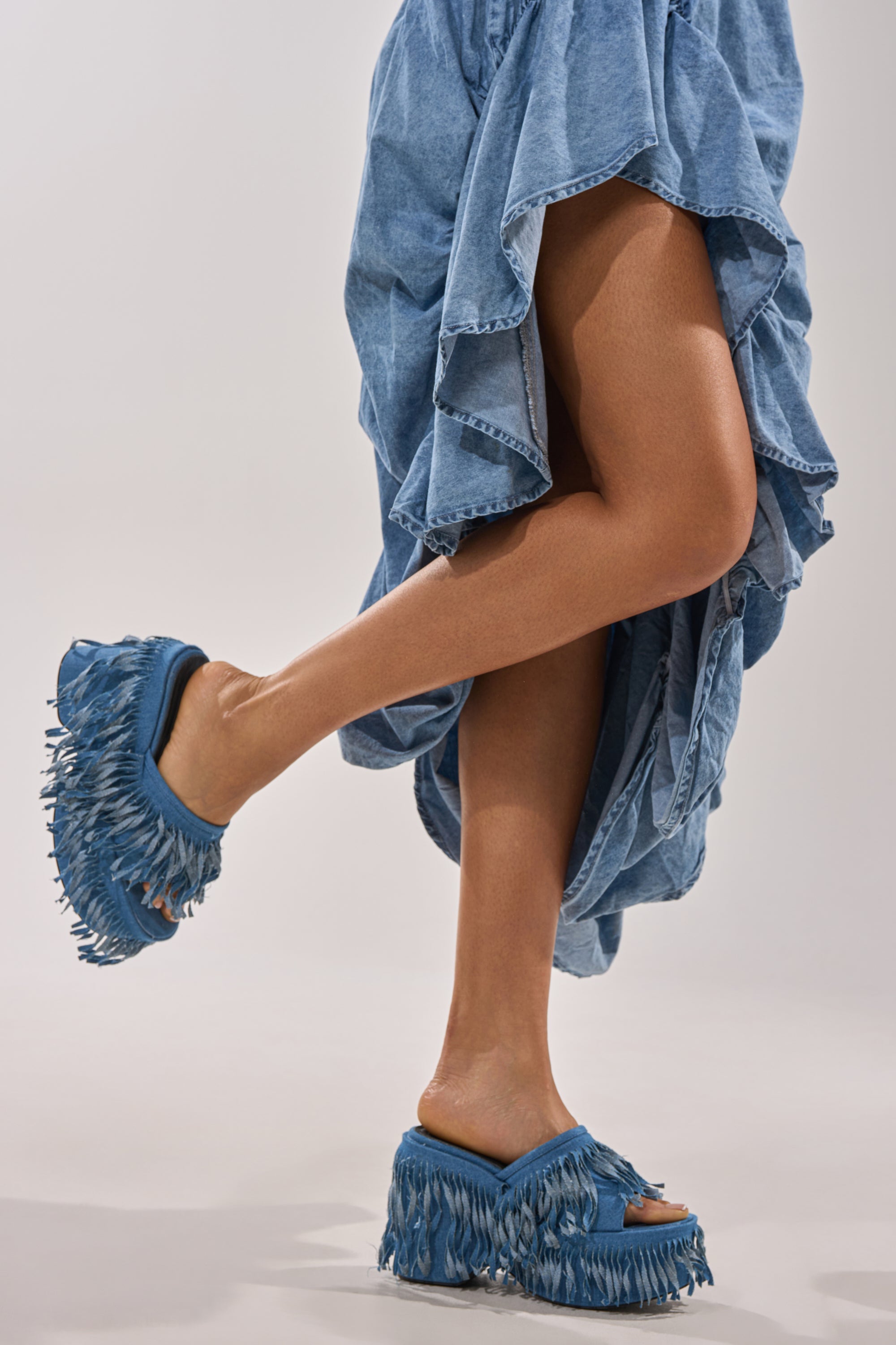 A person lifts one leg to showcase the AKIRA GOLD RAMESHA DENIM FRINGE PLATFORM SANDAL, styled with a ruffled high-low denim skirt, highlighting the unique footwear against a plain background. 1