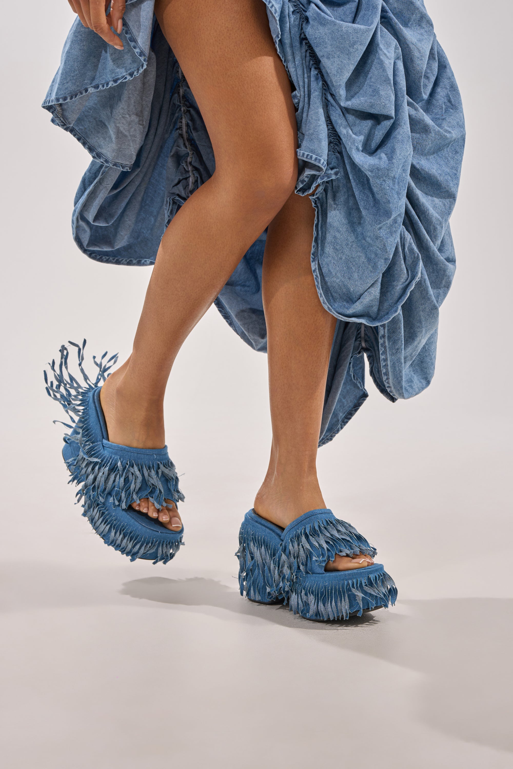 A person shows off their legs in the AKIRA GOLD RAMESHA DENIM FRINGE PLATFORM SANDAL, paired with a blue ruffled denim skirt. The chunky heeled sandals stand out against a simple, light background. 2