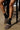 A person sitting on a stool sports the AZALEA WANG RAMIE LEOPARD MULE FLATS with buckle details and a faux fur upper, paired with black socks. They wear a black fringe garment against a plain brown floor backdrop.