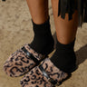 A person stands on a textured surface, featuring the AZALEA WANG RAMIE LEOPARD MULE FLAT slippers adorned with faux fur uppers and black straps over black socks. A fringe detail enhances the top of the image, adding a stylish touch.