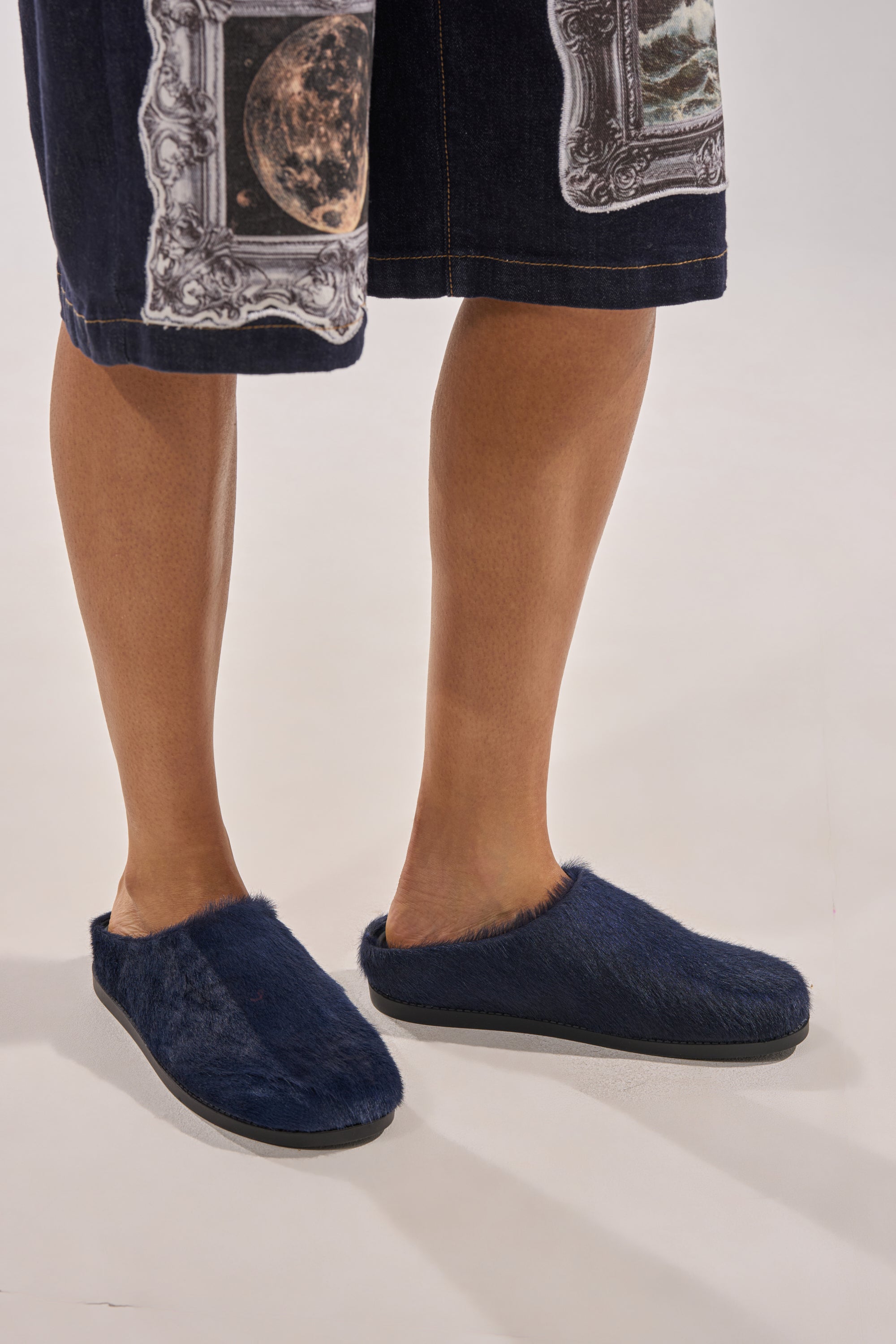Wearing the AZALEA WANG RAMSES NAVY SLIP ON FLAT, a person stands on a light background. Only their legs and feet are shown, paired with a dark celestial-printed skirt. The mules have a furry finish and slip-on style. 0