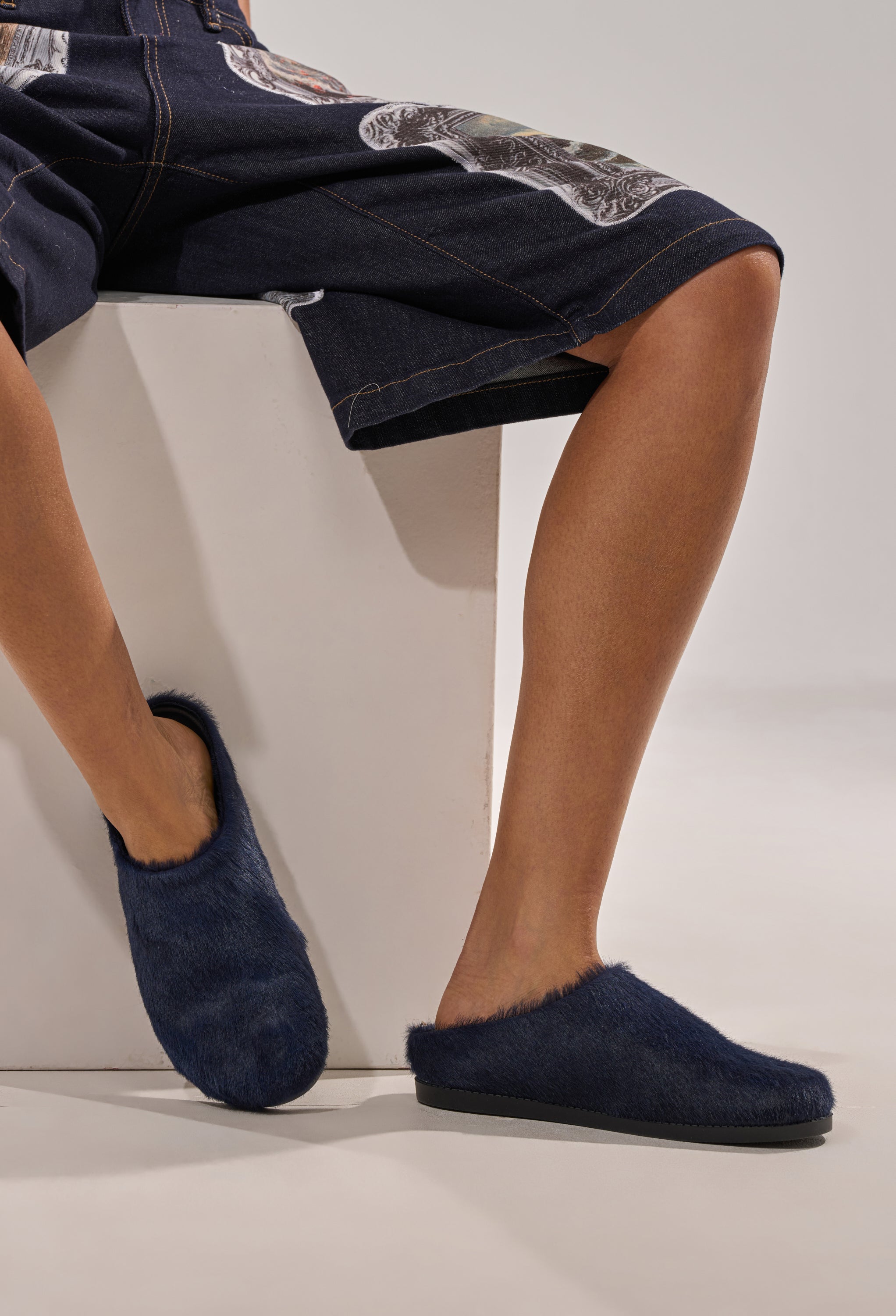 A person in dark patch shorts sits on a white block, displaying their bare legs and feet in AZALEA WANG RAMSES NAVY SLIP ON FLATS. 1