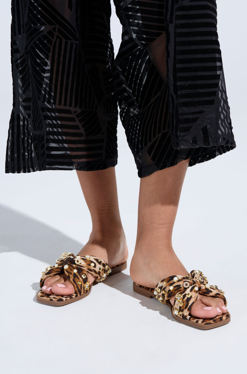 A person wearing black, sheer, patterned pants and AKIRA RAMSEY Leopard Flat Sandal with gold floral details in a comfortable slip-on fit against a light gray background.