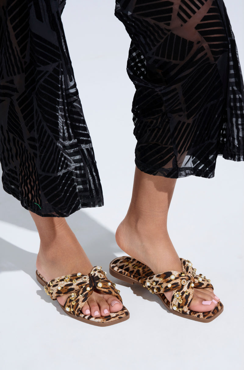 A person stands on a white surface wearing the AKIRA RAMSEY LEOPARD FLAT SANDALS, featuring embellished details. The slip-on design and cushioned insole ensure comfort while pairing beautifully with black, patterned sheer pants.