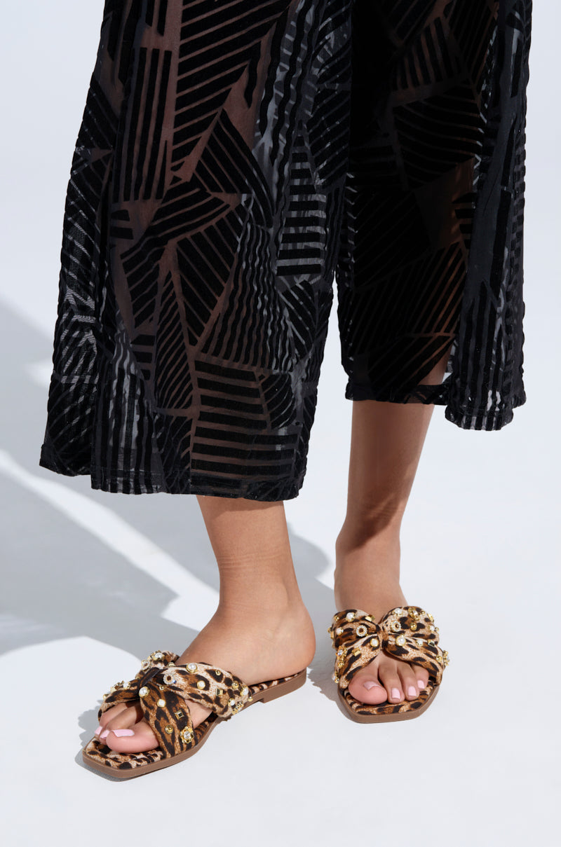A person is wearing black geometric-patterned pants and the AKIRA RAMSEY LEOPARD FLAT SANDAL, featuring a slip-on fit with a crisscrossed design embellished with beads and leopard print.