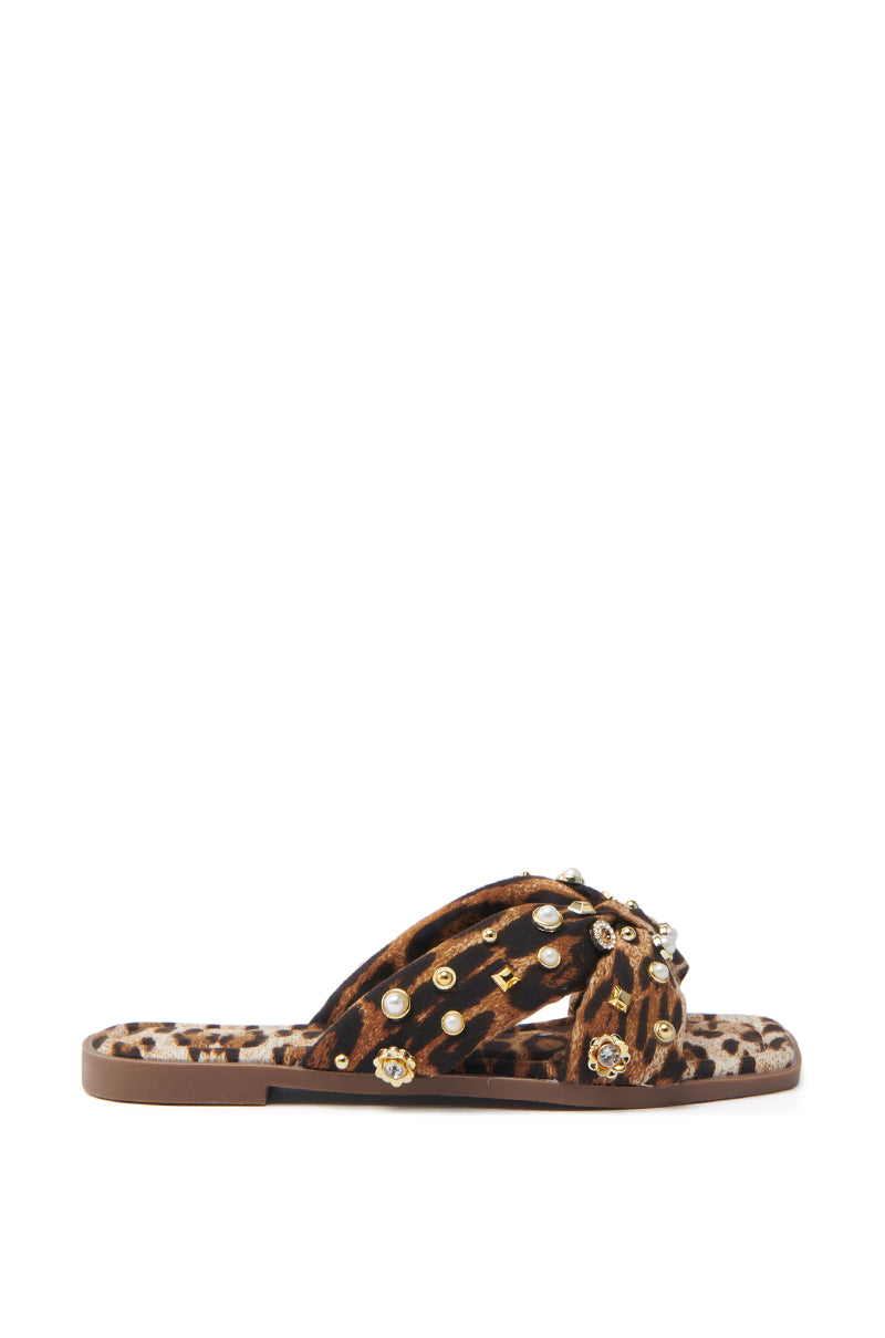 The AKIRA RAMSEY LEOPARD FLAT SANDAL offers a mix of comfort and style with its luxe leopard print and cozy flat sole. Featuring crisscross straps embellished with gold studs and circular gems, these sandals, set against a white background, promise flair in every step.