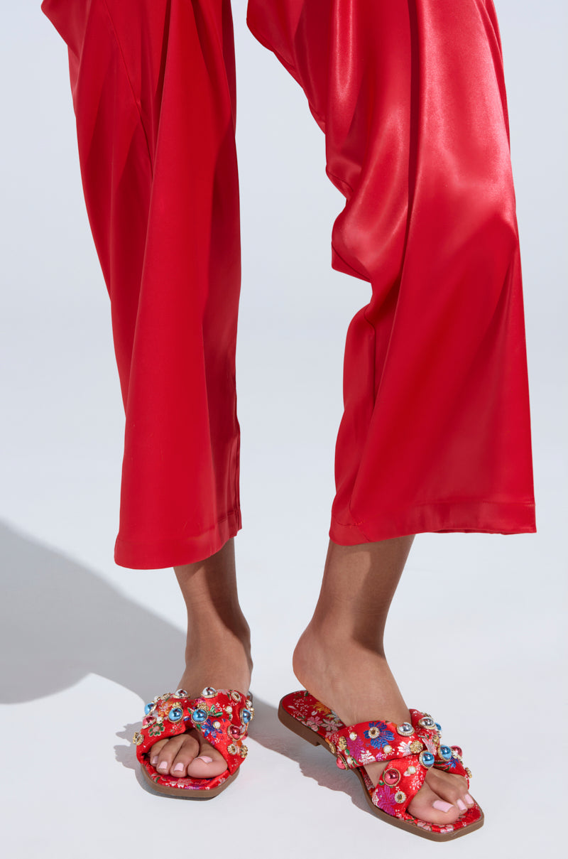 An individual wears shiny red trousers with AKIRA RAMSEY RED FLAT SANDAL, featuring vibrant floral embellishments, beads, and cushioned insoles on a light background.