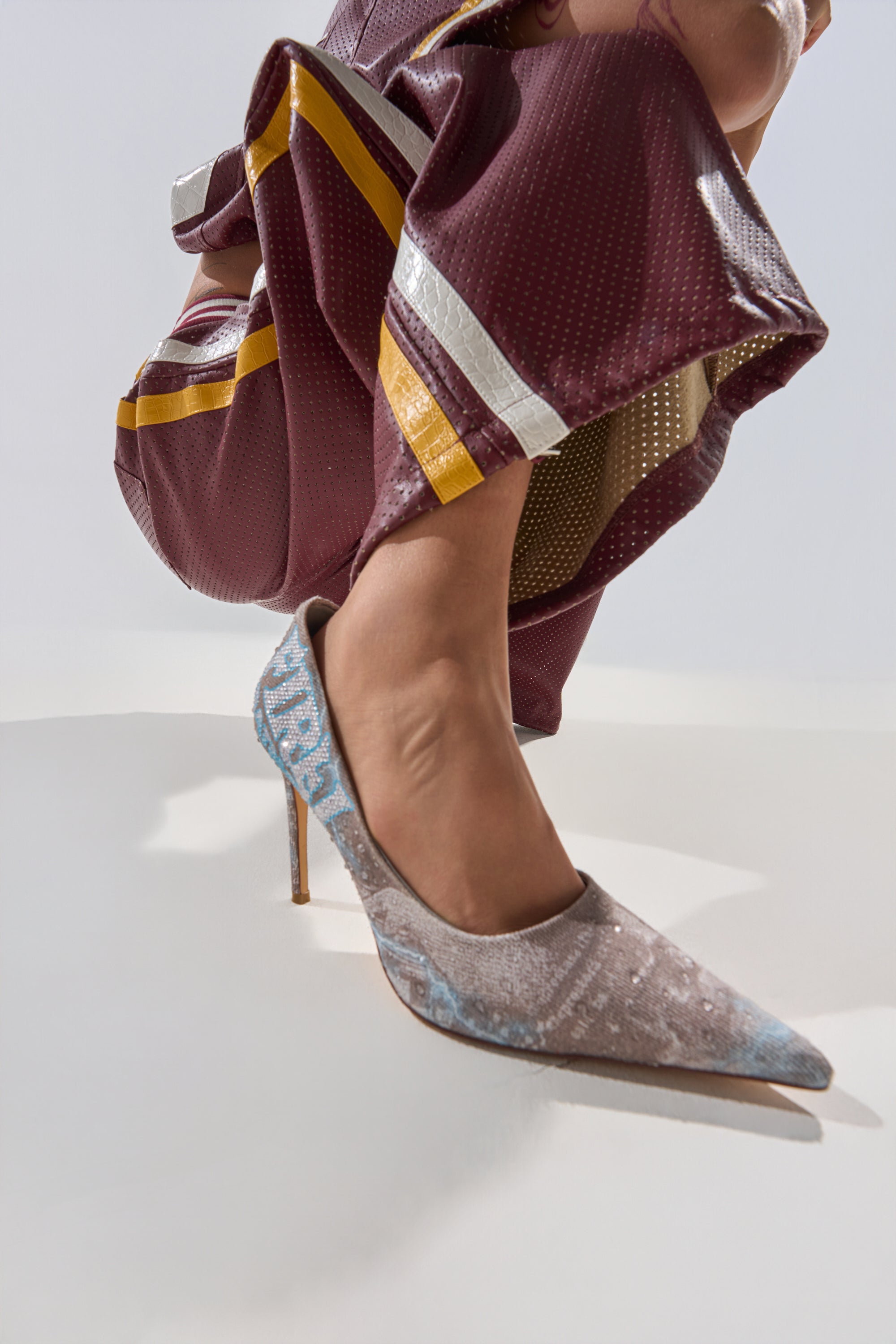 A person crouches in maroon pants with yellow and silver stripes, showcasing the AKIRA GOLD RANDA GREY MULTI PUMP—sparkly pointed-toe stiletto heels—in focus against a light background. 0