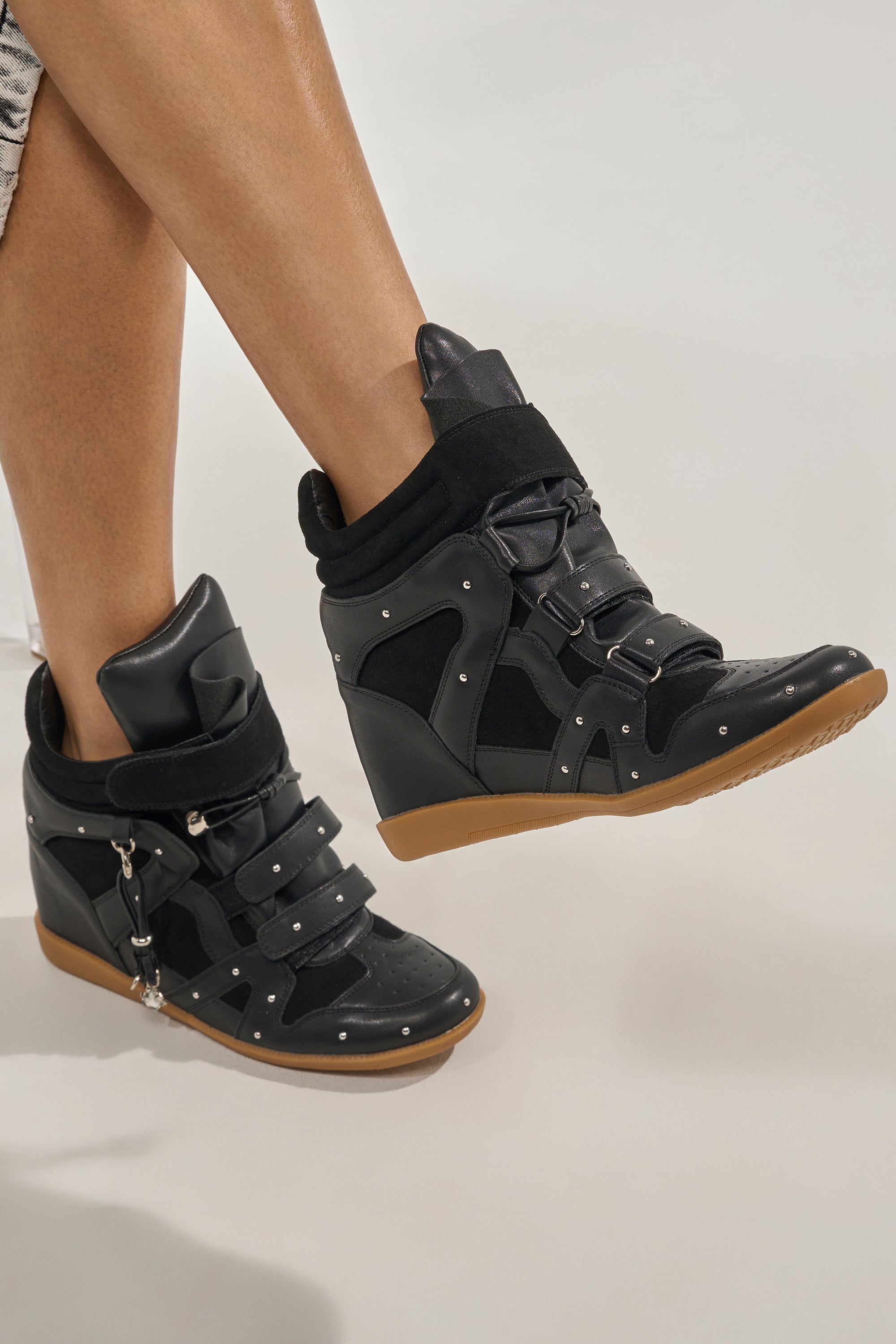 A person wears AKIRA GOLD RANGELEY black wedge sneakers with silver hardware, buckles, straps, studs, and a gum sole. Their legs are shown against a light background. 0