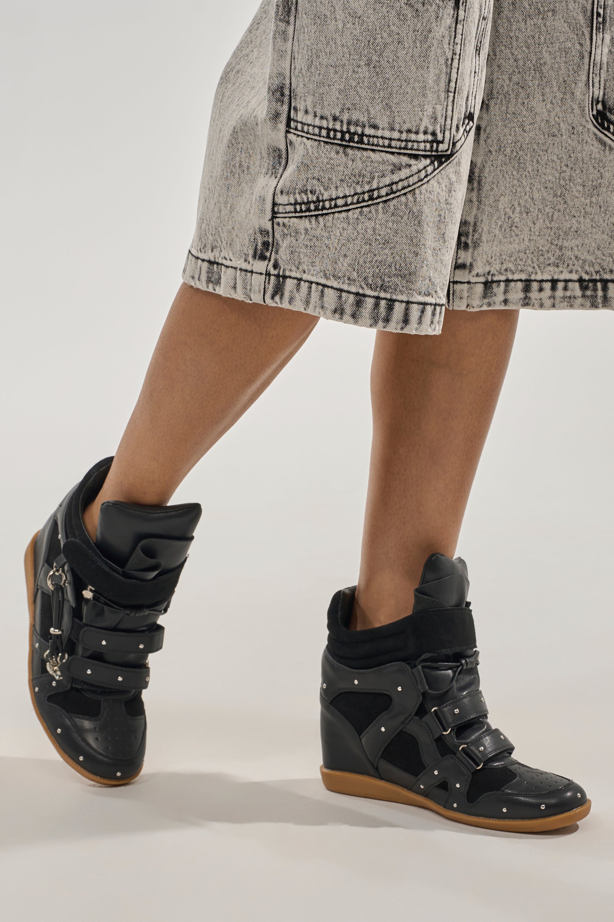 A person wearing a light-wash, knee-length denim skirt and AKIRA GOLD RANGELEY BLACK SILVER HARDWARE EMBELLISHED WEDGE SNEAKER stands on a neutral background. 7