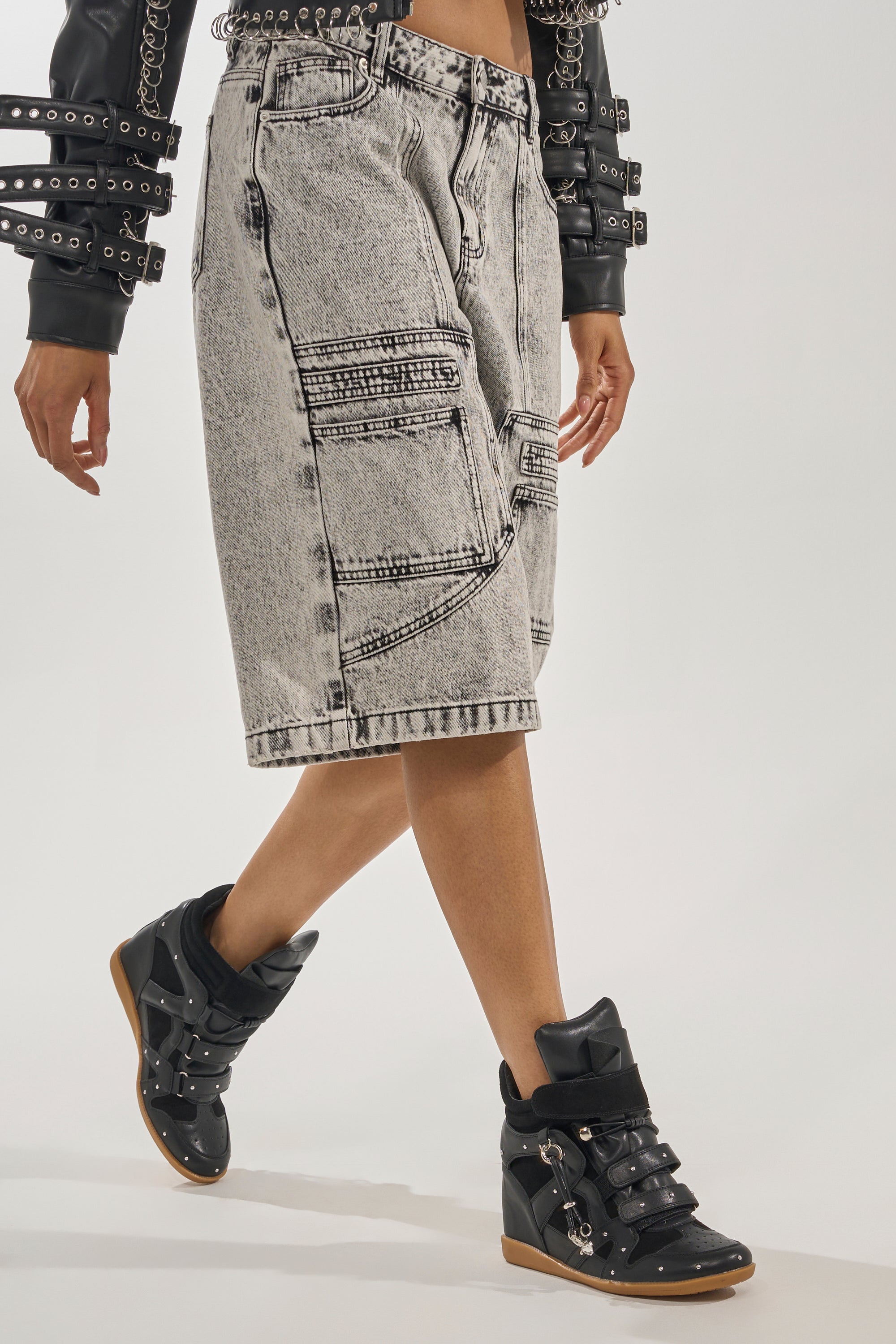 A person wears a black studded leather jacket, acid-wash knee-length denim shorts with multiple zippers, and AKIRA GOLD RANGELEY BLACK SILVER HARDWARE EMBELLISHED WEDGE SNEAKERS, walking against a plain background. 1