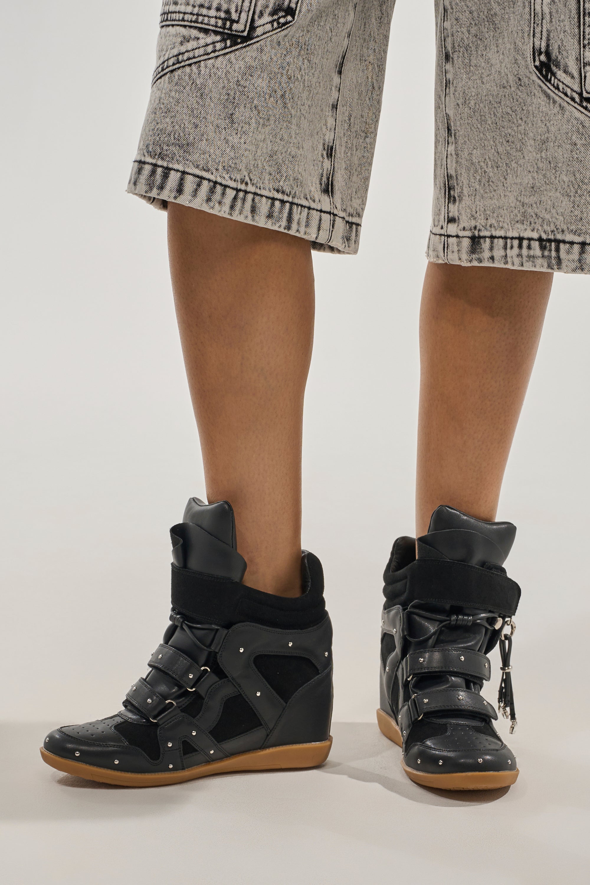 A person stands on a plain light background wearing light-wash, knee-length denim shorts and the AKIRA GOLD RANGELEY BLACK SILVER HARDWARE EMBELLISHED WEDGE SNEAKER. 6