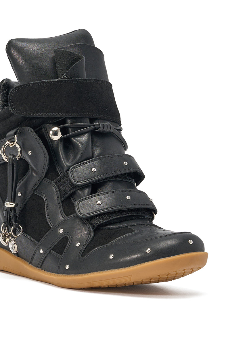 The AKIRA Gold Rangeley Black Silver Hardware Embellished Wedge Sneaker features black faux leather, velcro straps, silver studs, metal rings, and a light brown rubber sole shown close-up against a white background. 5