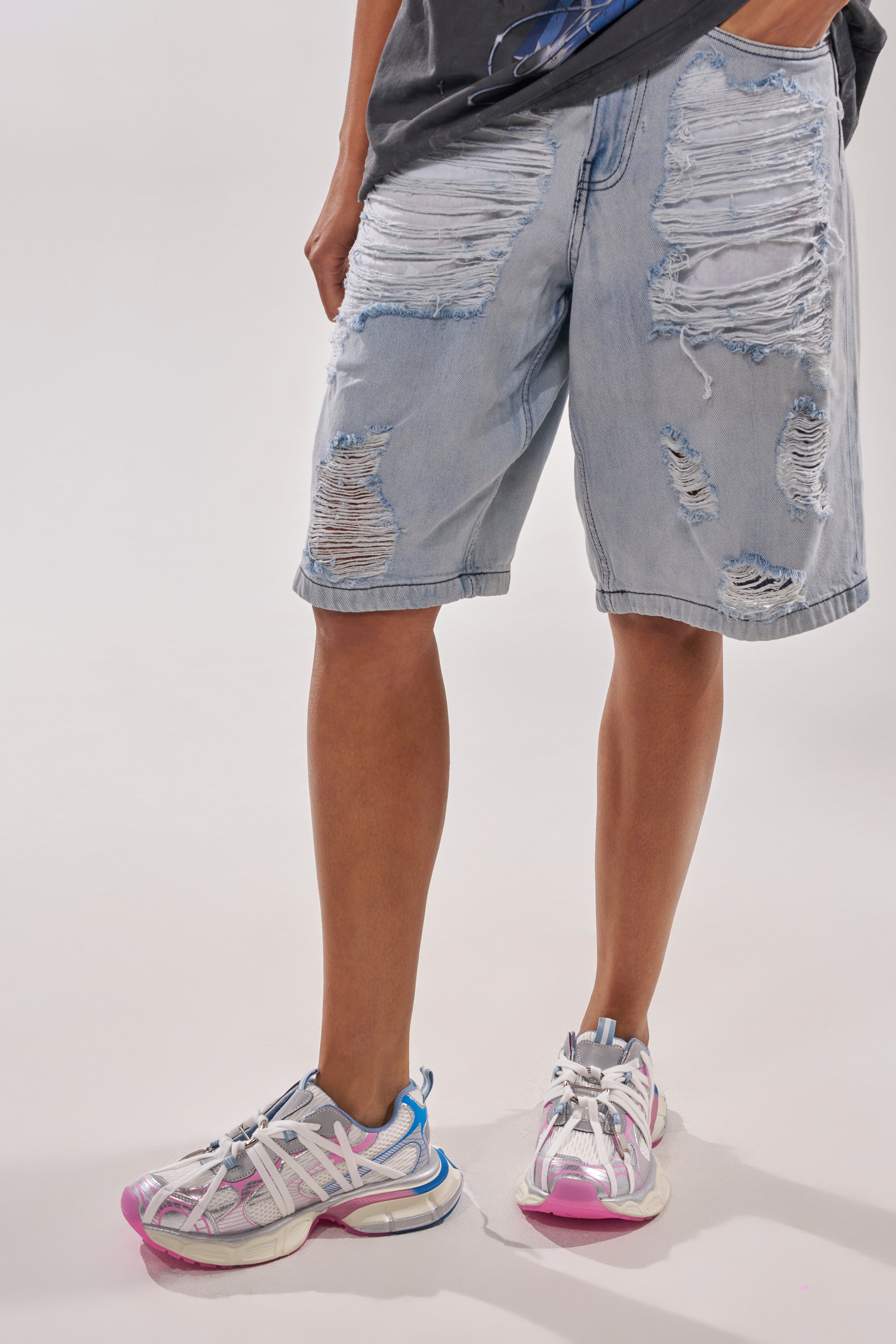 A person stands wearing light blue distressed denim shorts, a dark shirt, and AZALEA WANG RANGLE PINK MULTI LACE SNEAKERS with blue, silver, and pink details. The shorts feature large rips and frayed patches on the front. 0