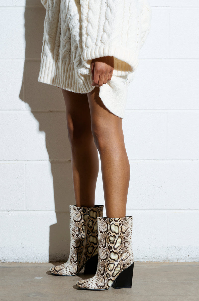 A person stands against a white brick wall, wearing a white cable-knit sweater dress and the chic AZALEA WANG RANSOM BLACK MULTI SNAKE BOOTIE featuring chunky heels.
