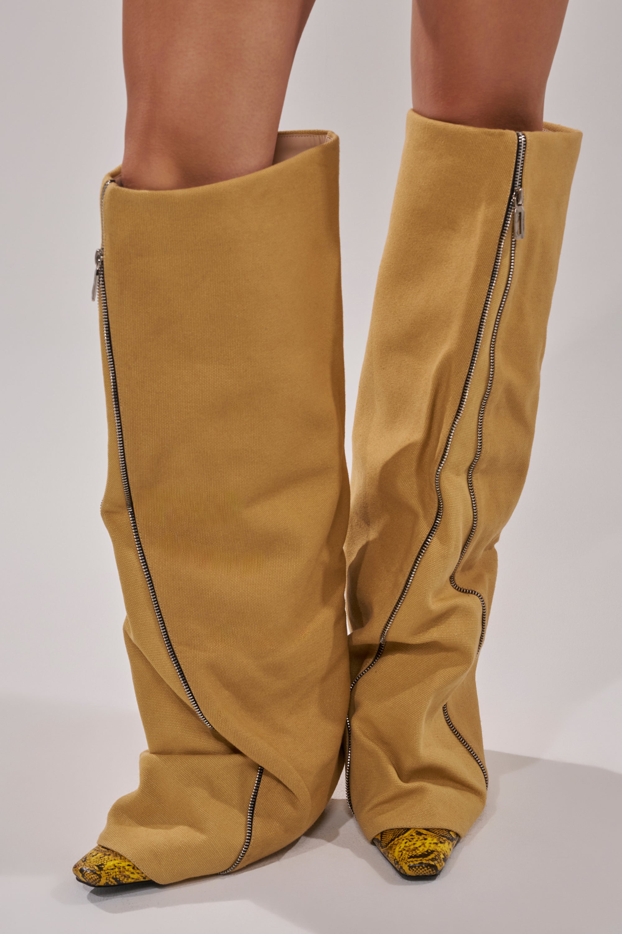 A person wears the AKIRA GOLD RASHAWN KHAKI AND SNAKE BOOT—oversized, slouchy over-the-knee boots featuring visible diagonal zippers and pointed tips that partly reveal yellow and black details.