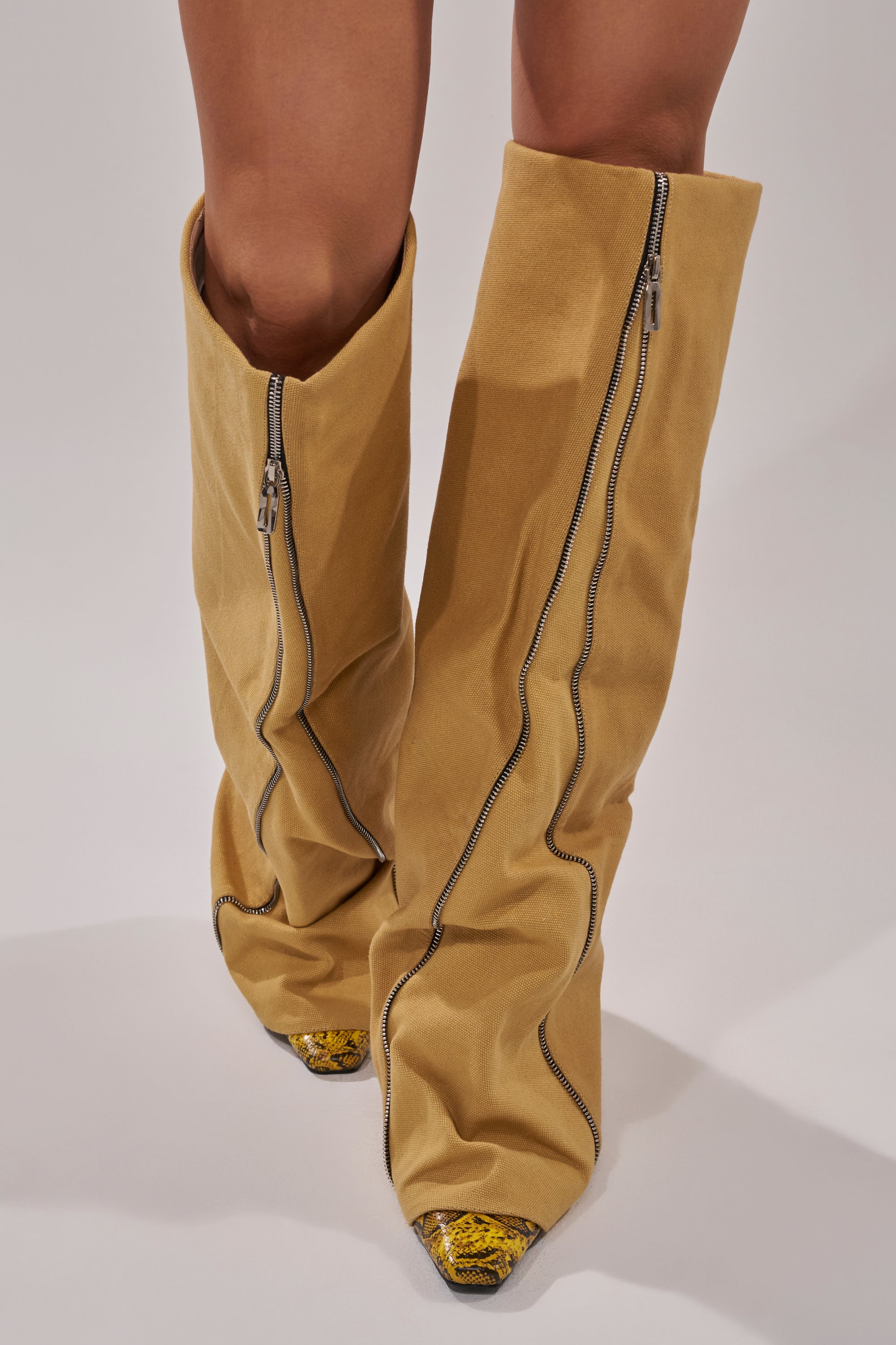 Close-up of a person wearing the AKIRA GOLD RASHAWN KHAKI AND SNAKE BOOT, featuring tall, loose khaki material, prominent front zippers, and pointed metallic toes, standing on a plain light surface.