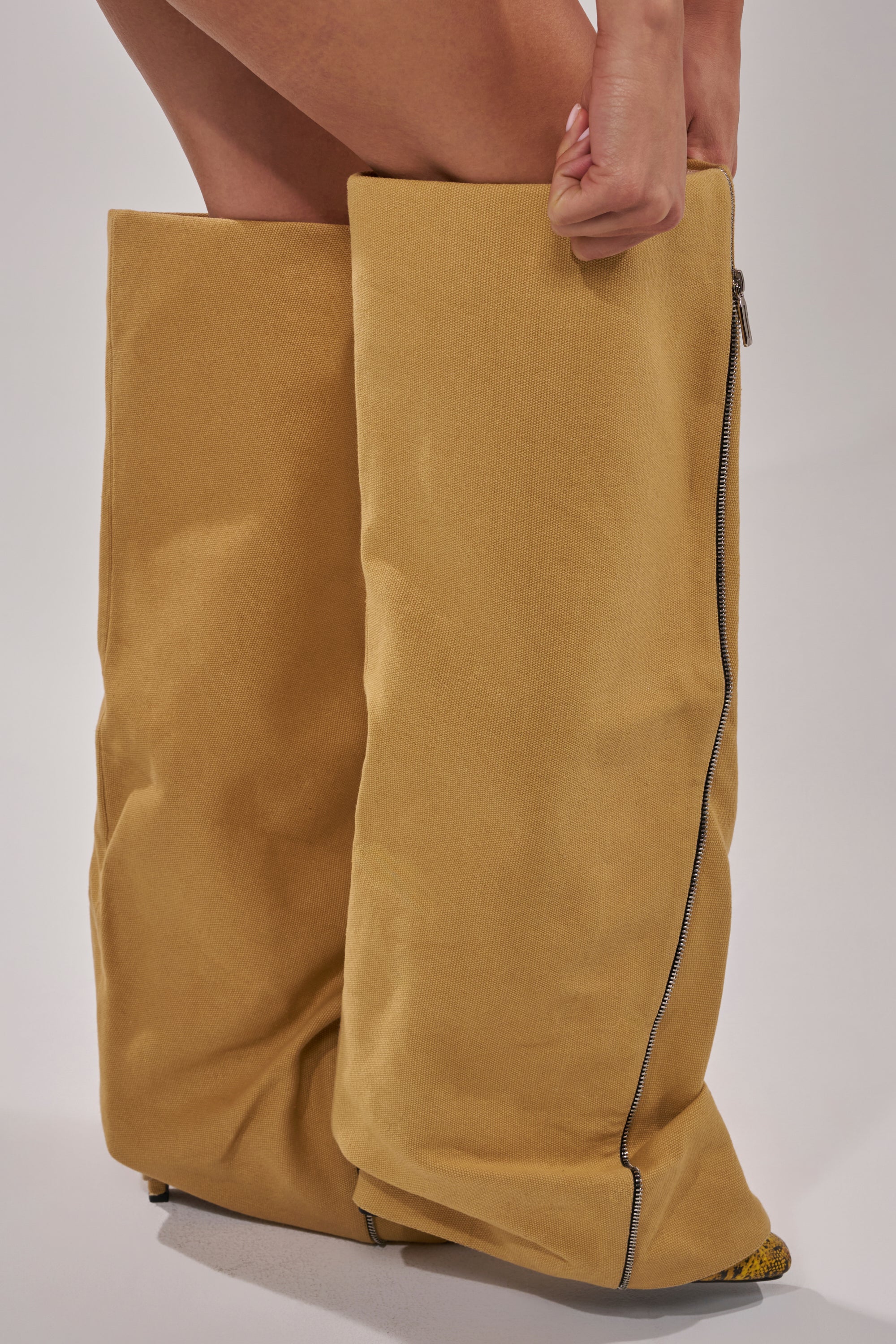 Someone pulls on the AKIRA GOLD RASHAWN KHAKI AND SNAKE BOOT—oversized, baggy, tan faux leather boots with side zippers that extend over the knees. The scene features a plain, light background.