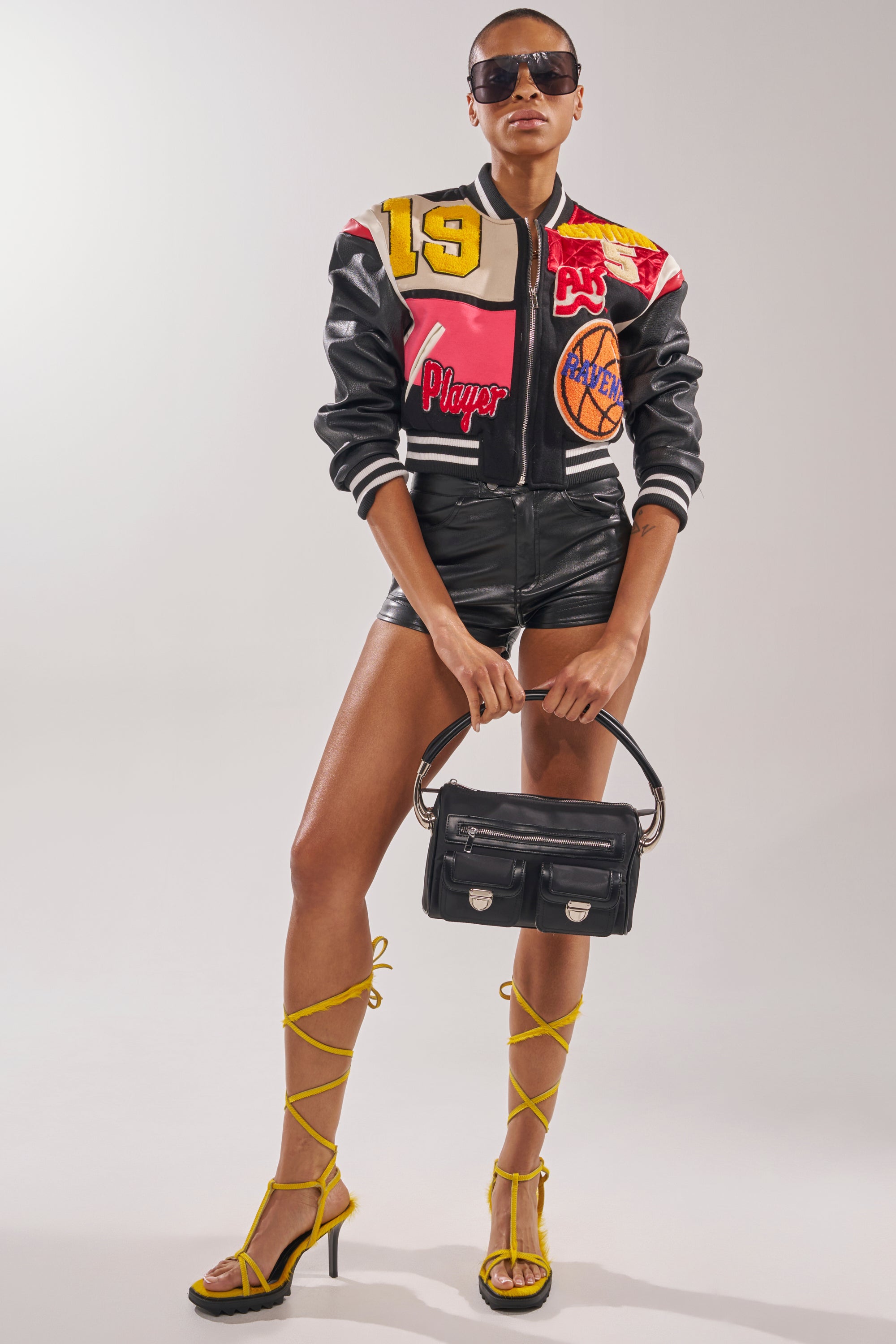 A person poses confidently against a light background, wearing colorful attire and accessorizing with the AKIRA GOLD RAVZA YELLOW CALF HAIR STRAP HIGH HEEL STILETTO SANDAL, large sunglasses, and a black handbag. 2