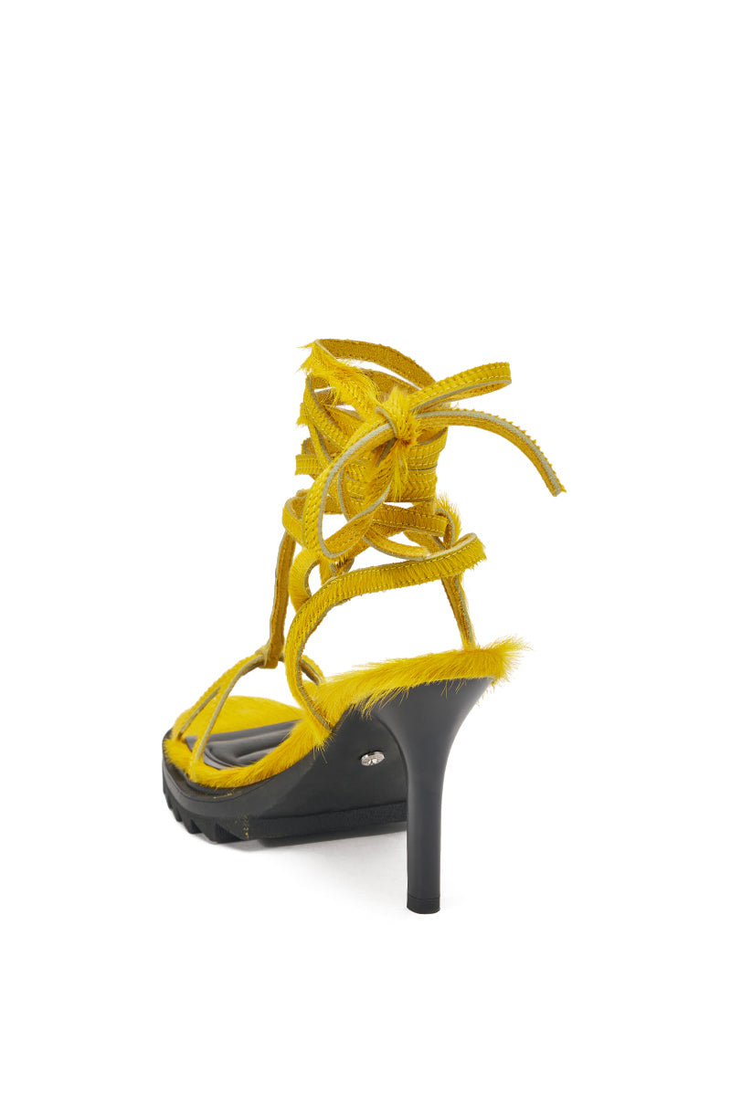 The AKIRA GOLD RAVZA YELLOW CALF HAIR STRAP HIGH HEEL STILETTO SANDAL features fuzzy yellow calf hair straps, lace-up ankle cords, an open toe, and a contrasting black heel and sole, shown from a rear side angle against a white background. 5