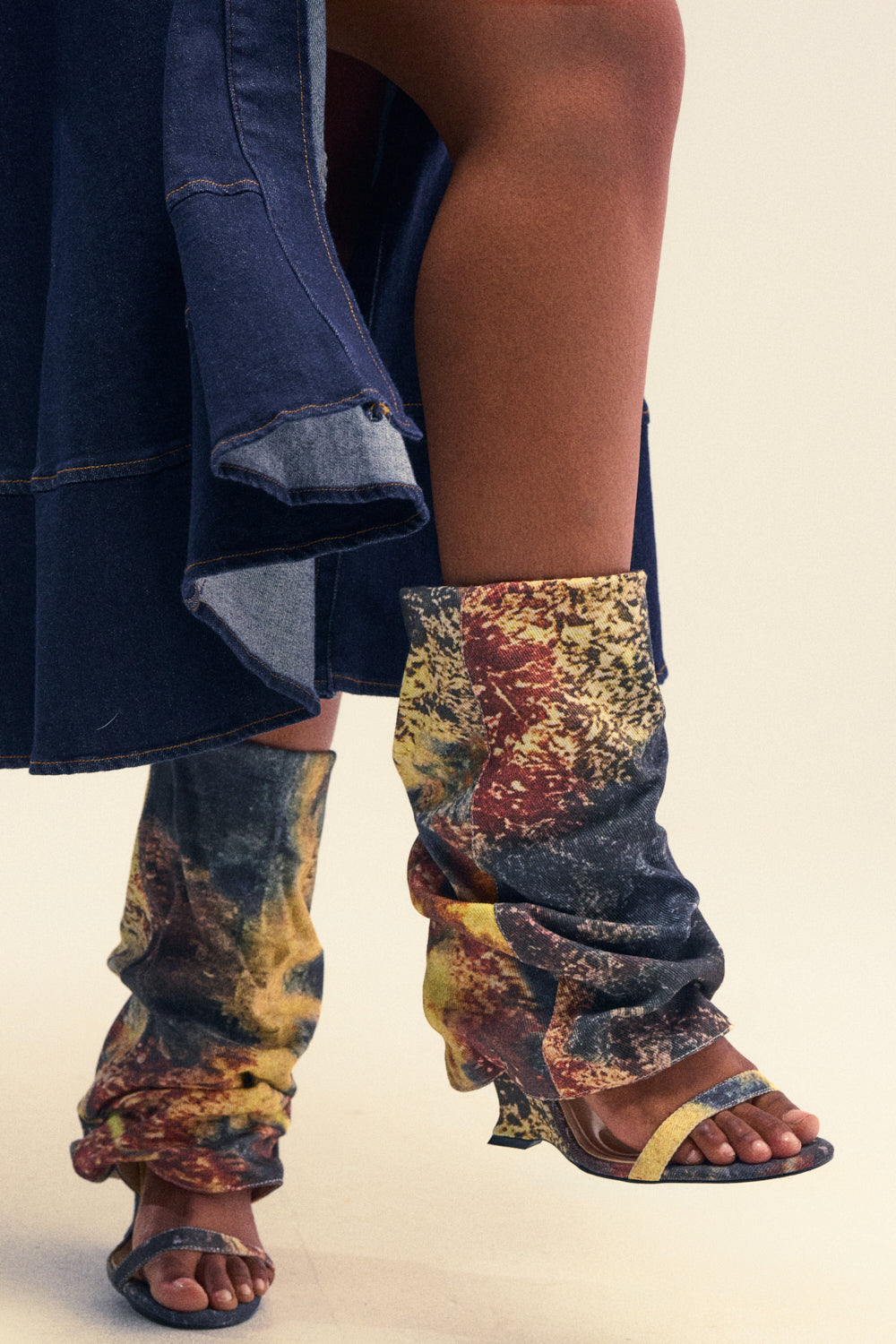 Wearing the AZALEA WANG RAXA MULTI OPEN TOE BOOTIE—colorful, slouchy mid-calf boots featuring abstract patterns and open toes and heels—and a denim skirt with a frayed hem, a person stands against a plain background.