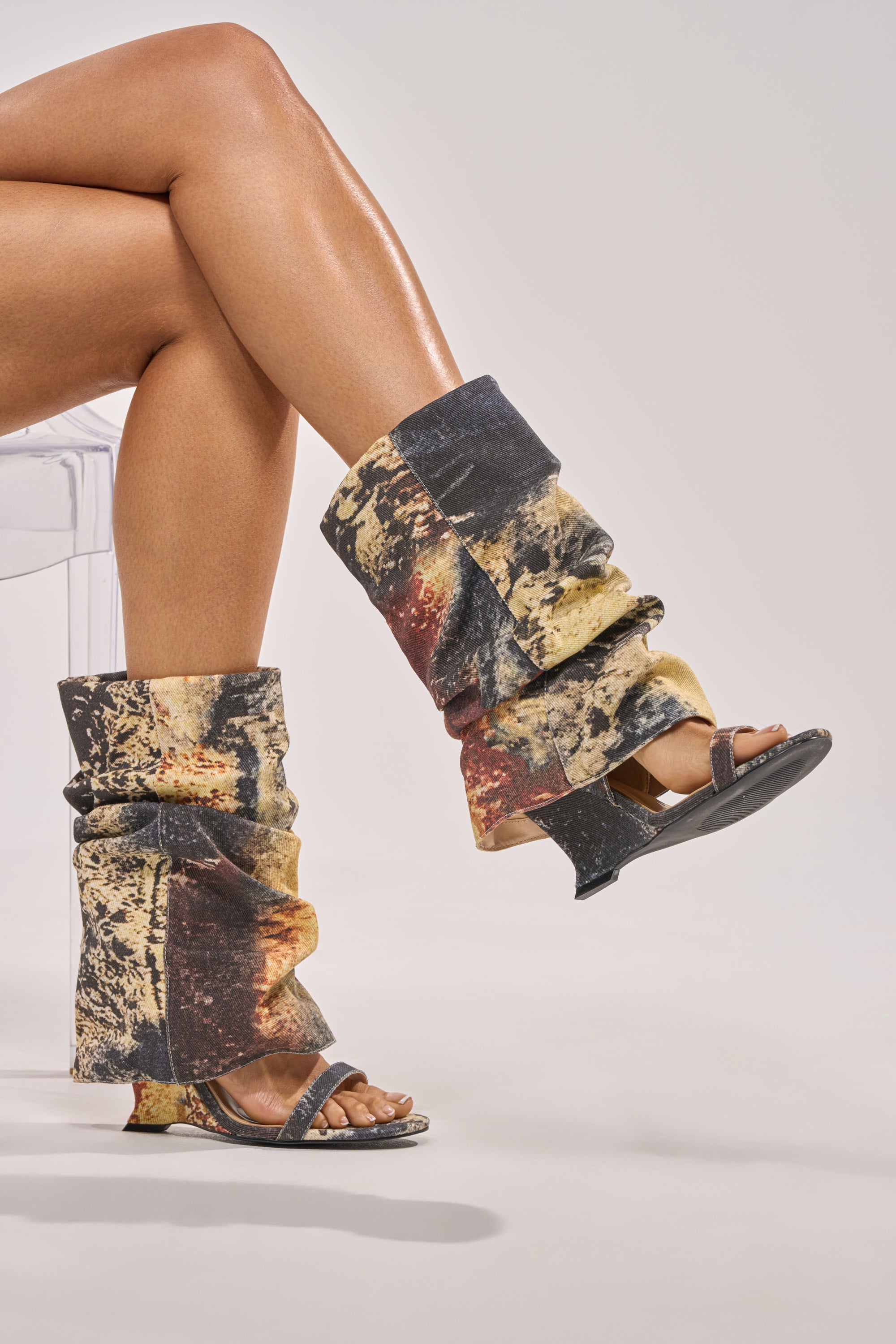 A person with crossed legs wears AZALEA WANG RAXA MULTI OPEN TOE BOOTIE and tall, multicolored abstract leg warmers. They sit on a transparent chair against a plain light gray background. 1