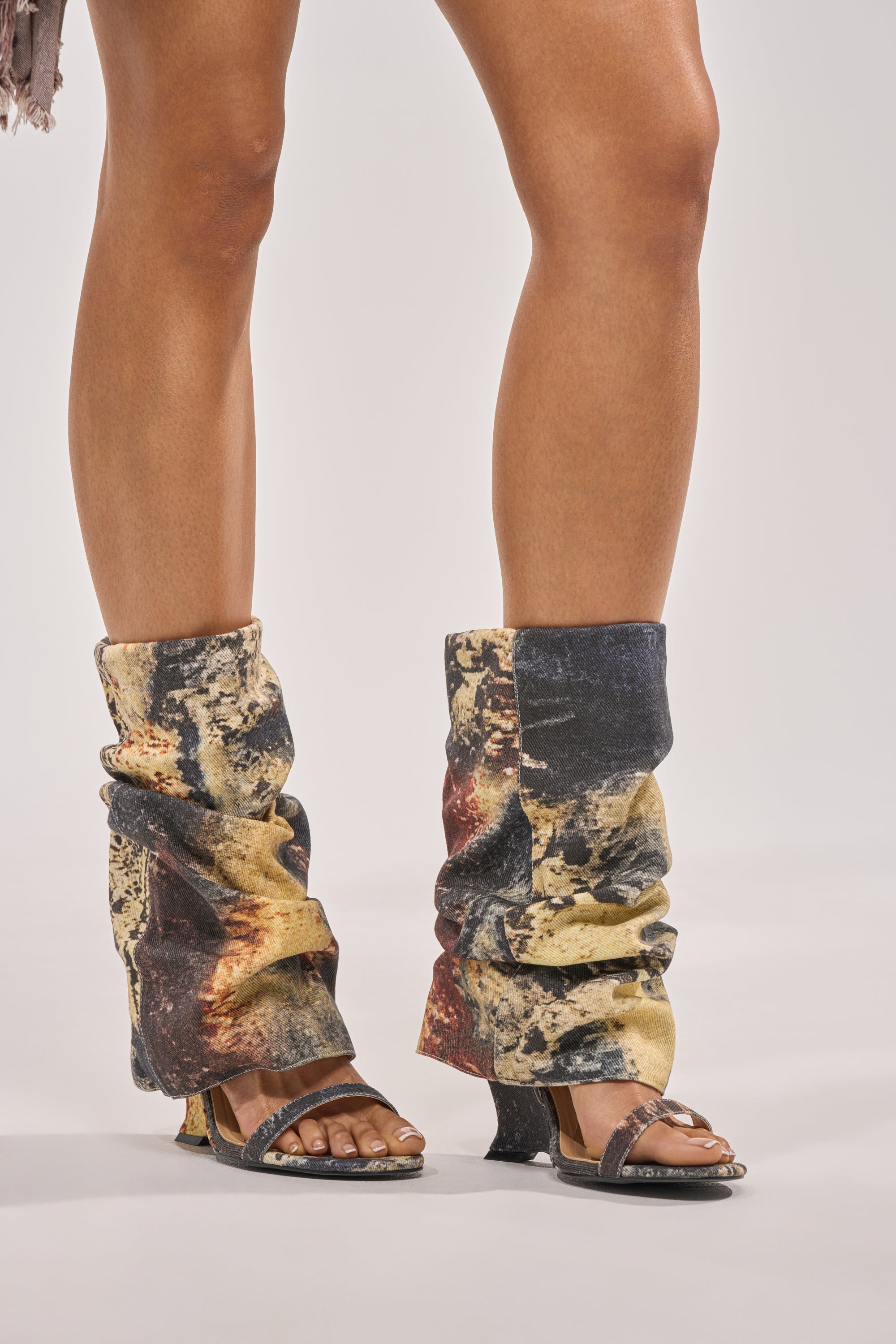 Wearing the AZALEA WANG RAXA MULTI OPEN TOE BOOTIE and slouchy leg warmers with an abstract yellow, black, and brown splatter paint pattern, a person stands on a white background. 2
