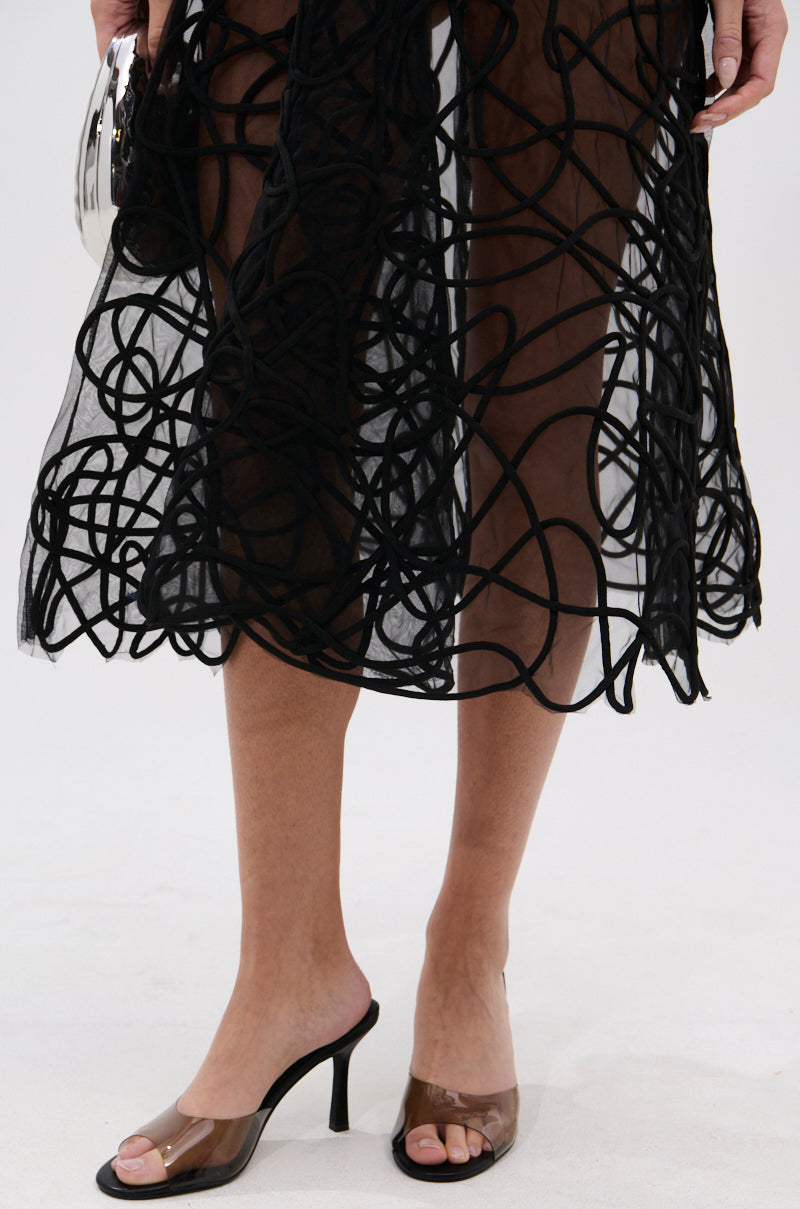 A person wearing a sheer black skirt with a swirling line design and the AKIRA RAYA BLACK PVC CHUNKY HEEL SANDAL, holding a small metallic handbag, stands against a plain light background.