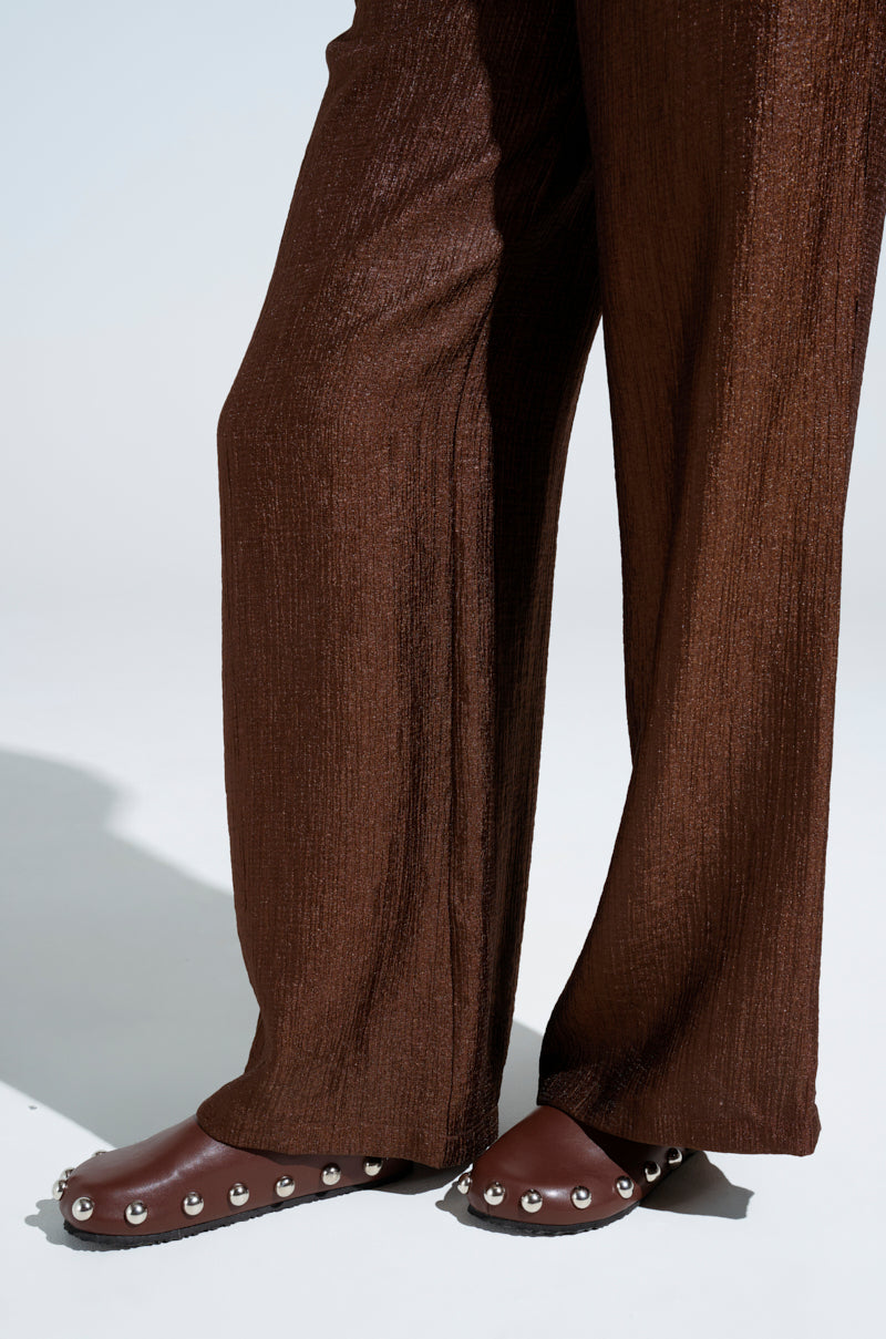 A close-up shows a person wearing brown textured wide-leg pants and AZALEA WANG RAYNE BROWN FLATS with almond toes, enhanced by metallic silver embellishments. The backdrop is a plain light color.