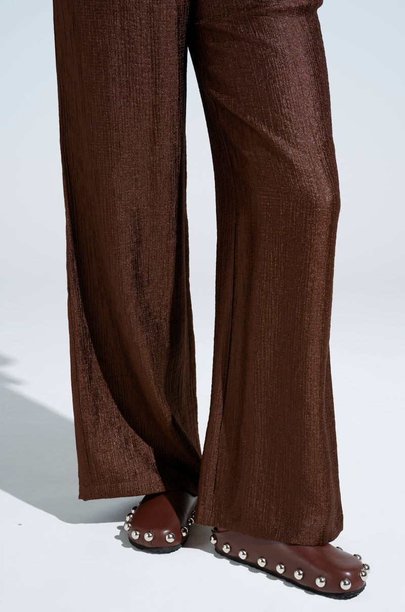 A person stands on a light gray background, dressed in brown textured wide-leg pants, paired with AZALEA WANG RAYNE Brown flats featuring metallic silver embellishments and an almond toe.