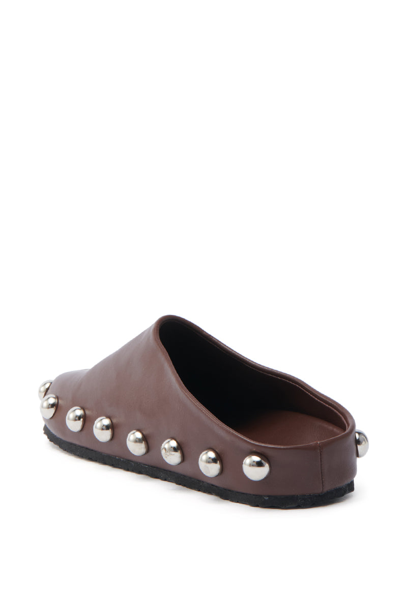 The AZALEA WANG RAYNE BROWN FLAT is a brown slip-on shoe with silver metallic studs on the edge, heel, and front. Made from vegan leather, it features a black flat sole, soft almond toe design, and an open back for easy access.