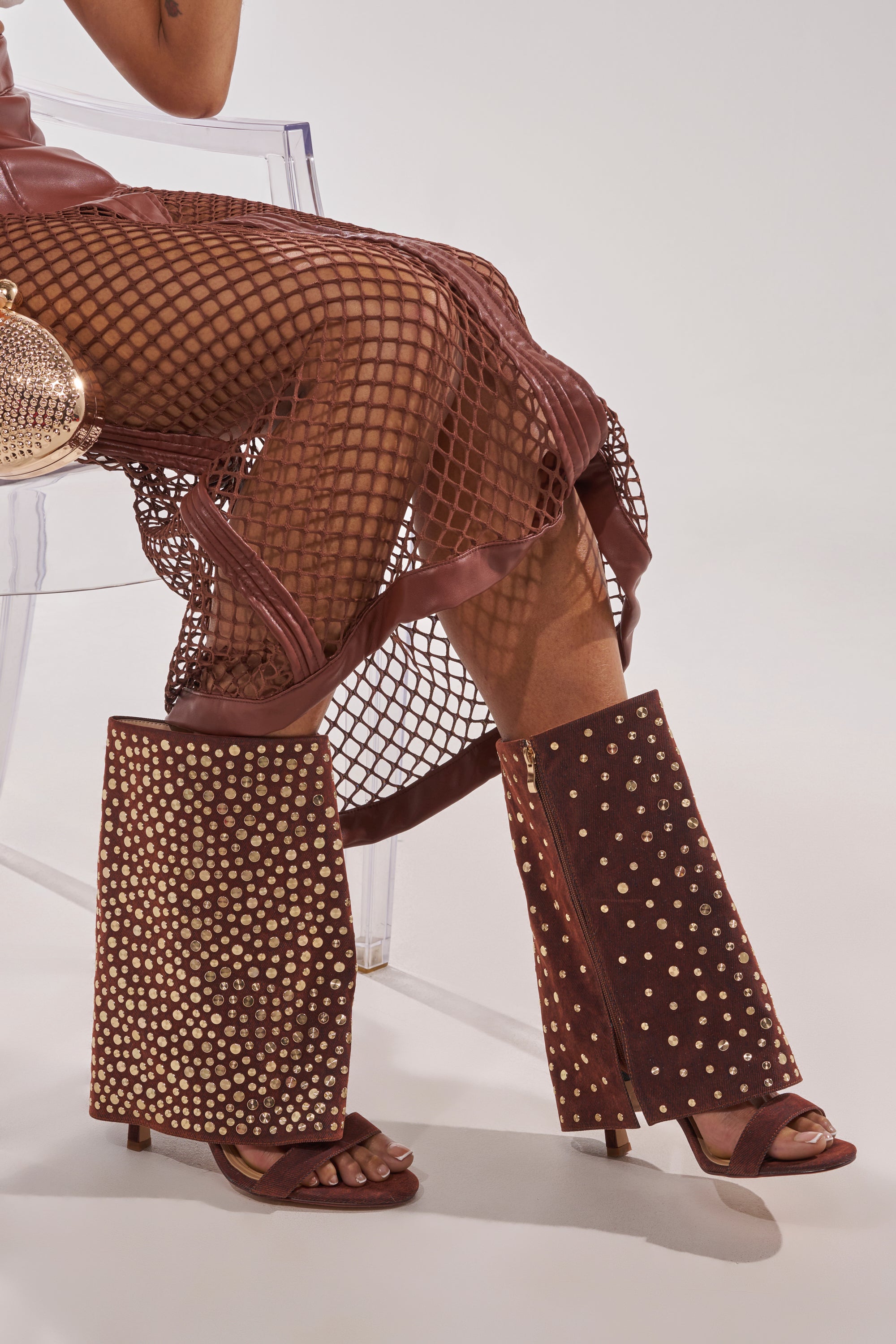 Seated on a clear chair, the person wears a brown fishnet skirt, unique brown chaps with gold studs over the AKIRA GOLD RAYSSA BURNT ORANGE GOLD HARDWARE EMBELLISHED STILETTO SANDAL, styled with a metallic mesh purse. 2