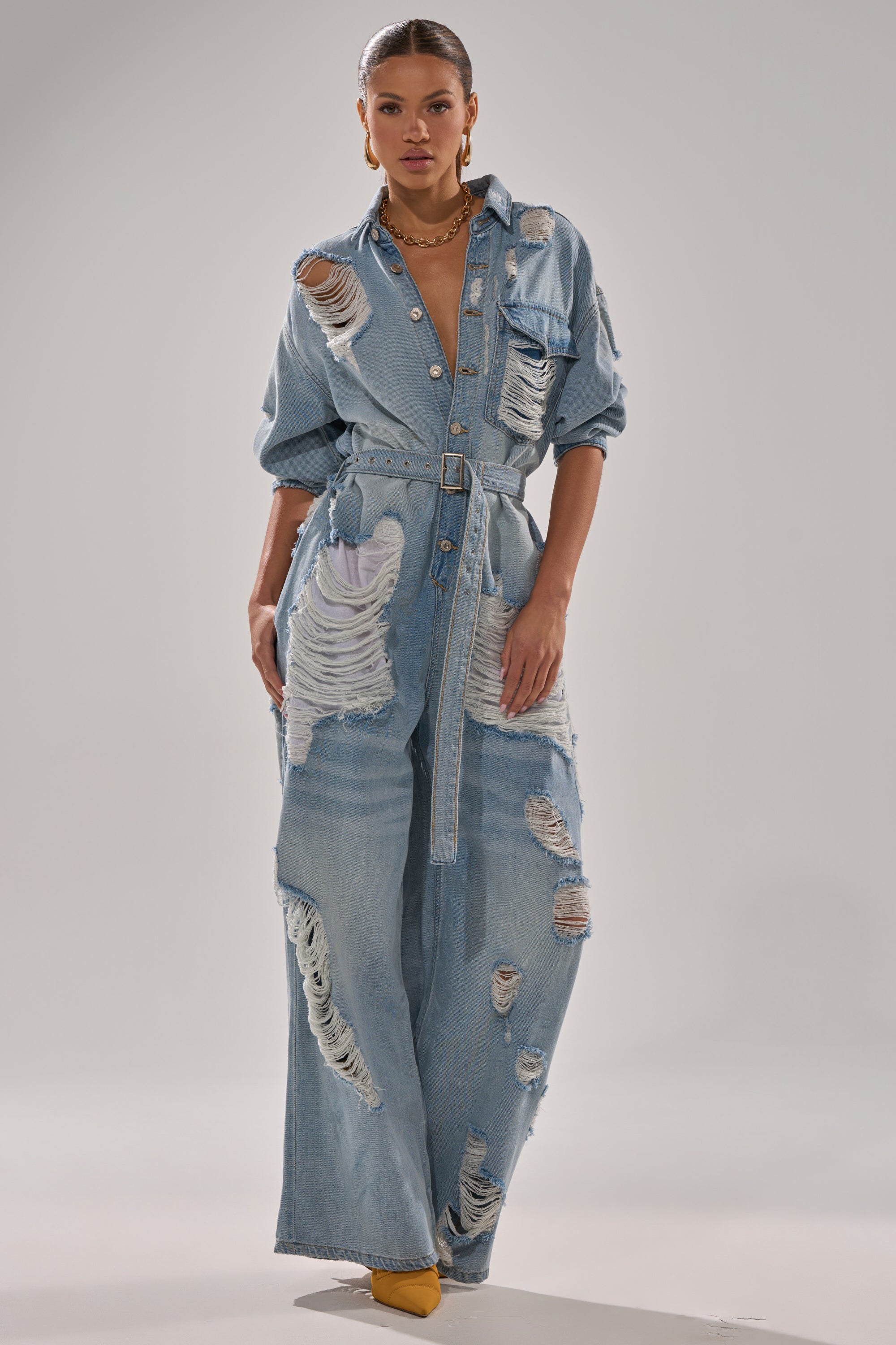 A woman stands confidently in the NOT TODAY EXTREME OVERSIZED DISTRESSED DENIM JUMPSUIT, featuring a light blue wash, belted waist, ripped details, and wide-leg fit. She accessorizes simply and pairs it with yellow pointed-toe shoes against a gray backdrop.