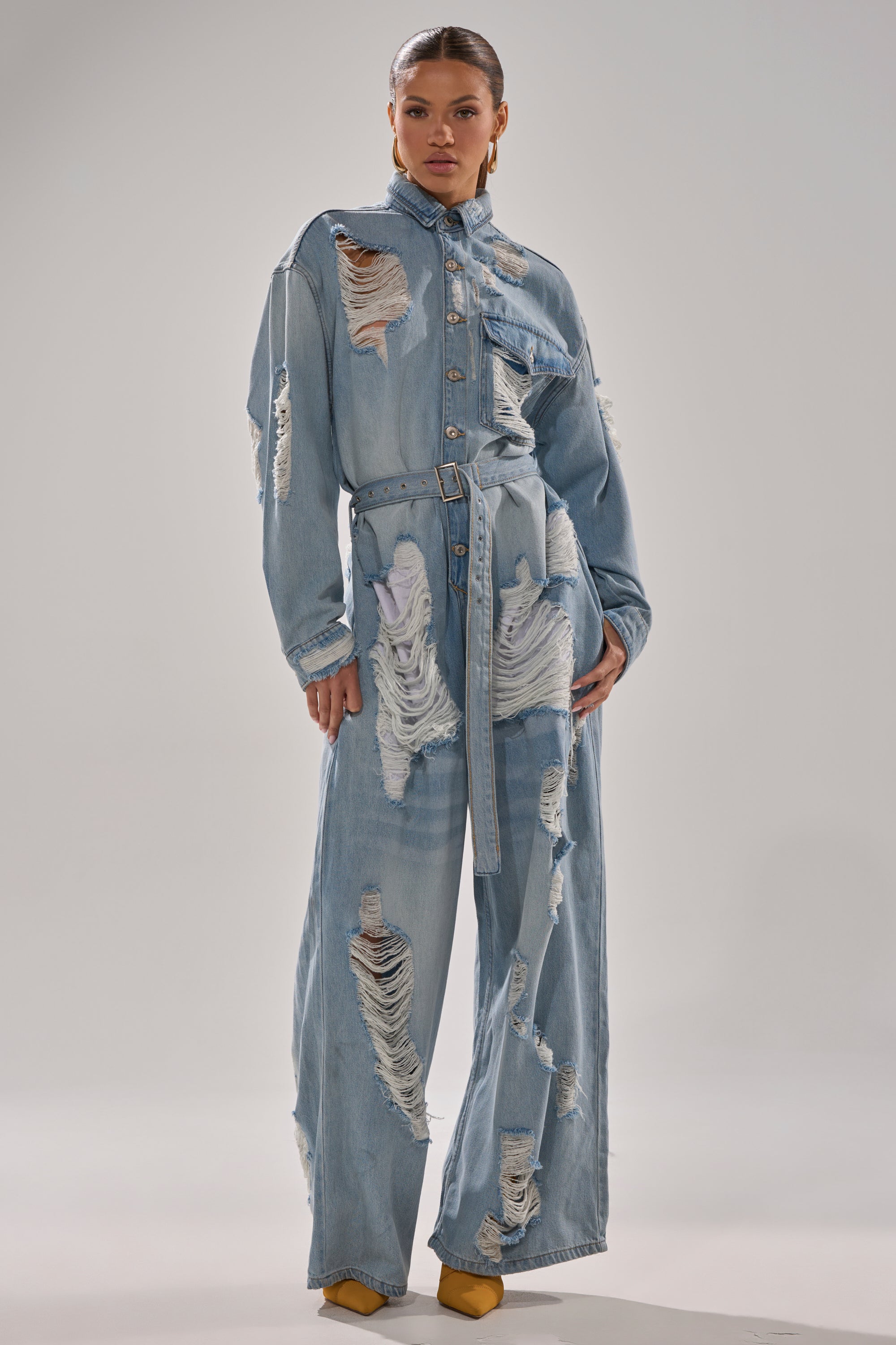 A person stands confidently in the NOT TODAY EXTREME OVERSIZED DISTRESSED DENIM JUMPSUIT with wide legs and heavy rips, paired with yellow pointed-toe shoes, against a plain background.