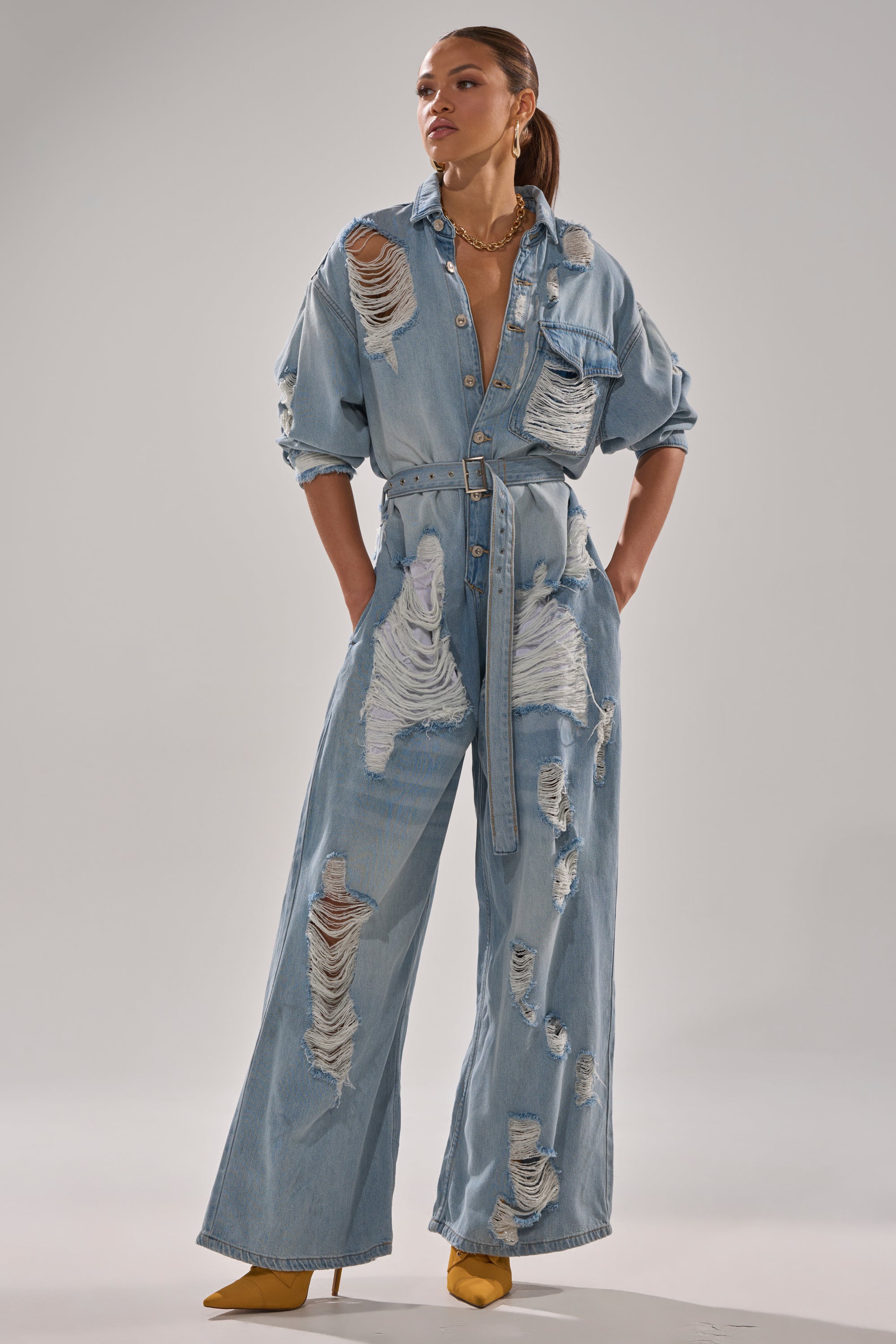 A woman models the NOT TODAY EXTREME OVERSIZED DISTRESSED DENIM JUMPSUIT, featuring a belted waist and ripped details, styled with yellow pointed-toe heels for a modern look against a plain background.